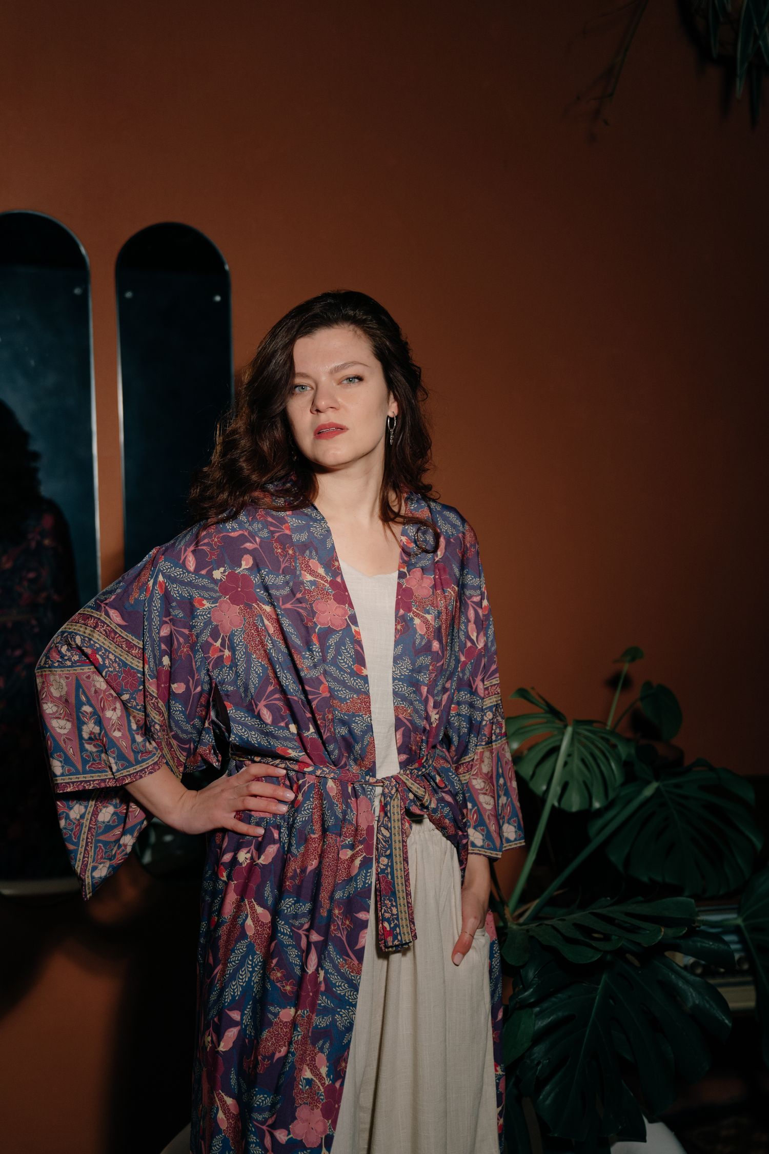 Woman in floral kimono robe poses confidently indoors with tropical plants and warm terracotta walls.