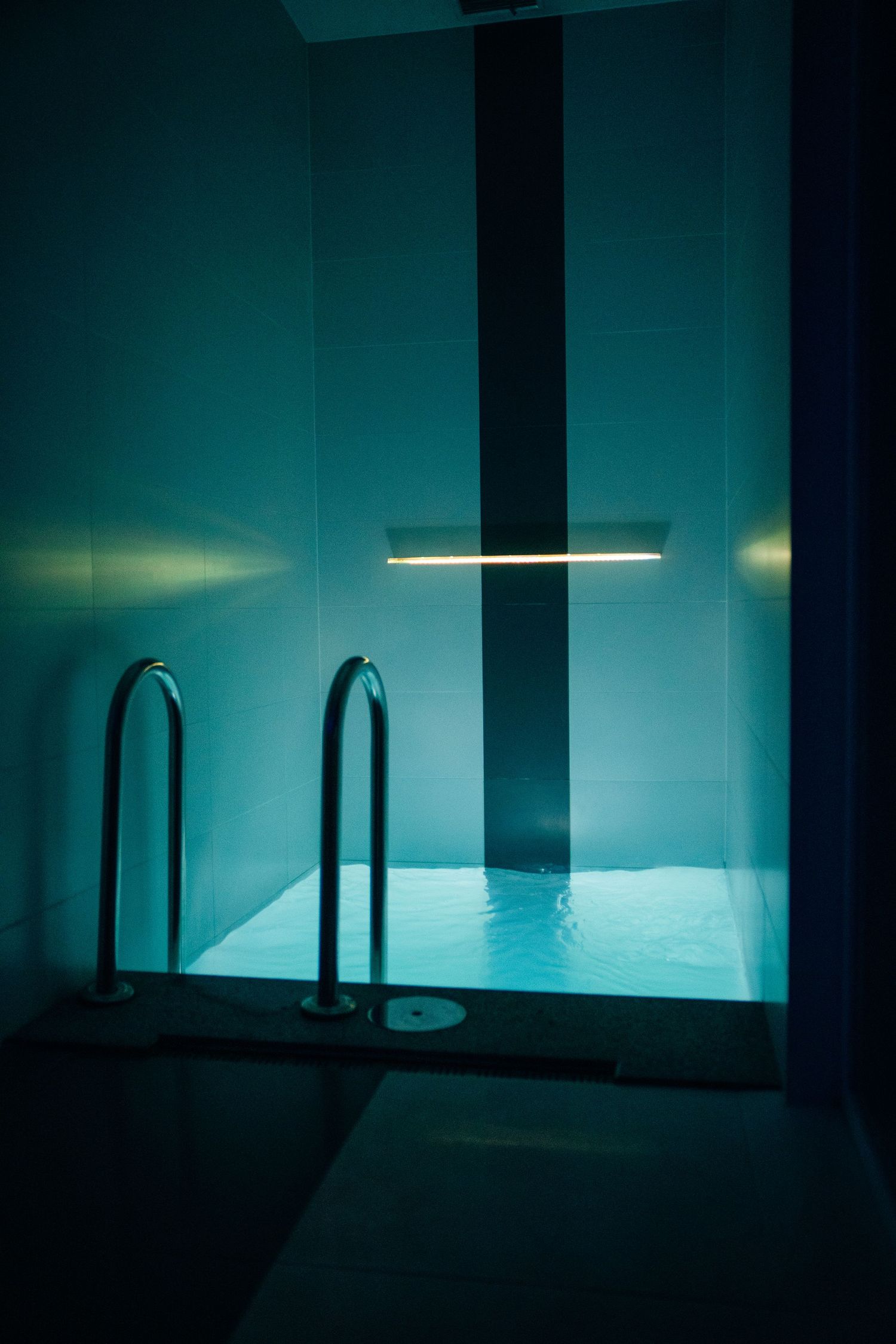 Moody indoor pool with teal lighting, metal handrails, and a glowing linear light fixture on the wall.