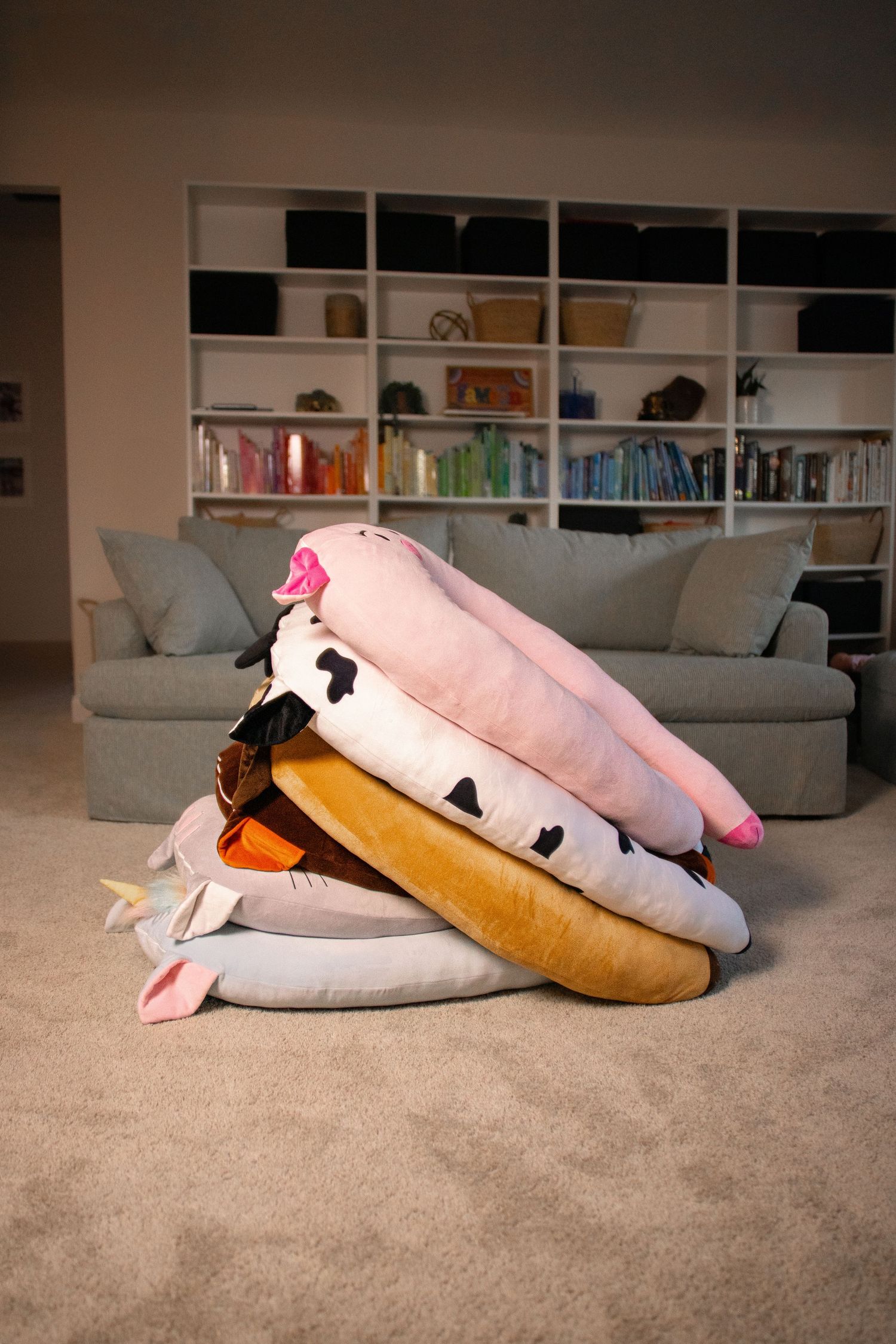 Stack of large animal-shaped plush pillows piled on carpet in cozy living room with bookshelf.