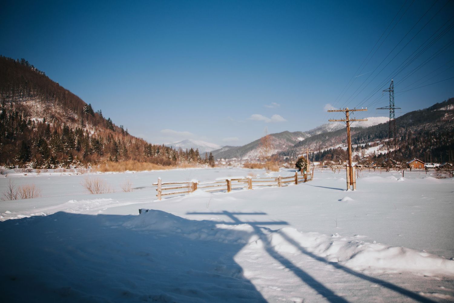 Snow-covered valley with wooden fence, power lines, and pine-forested mountains under a clear blue winter sky.