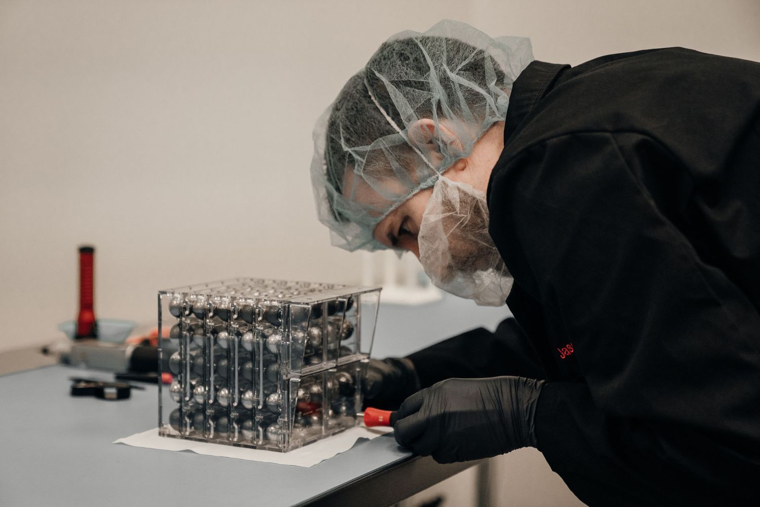 A technician in cleanroom gear carefully assembles an electronic circuit cube on a white workbench.