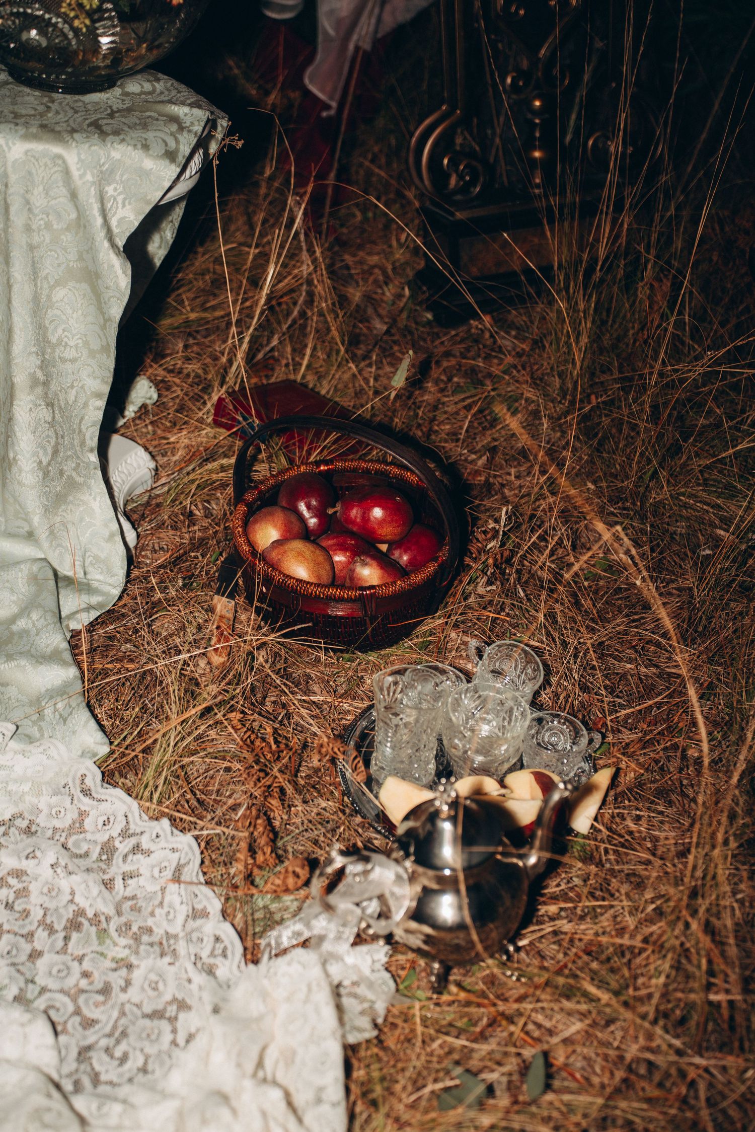 Rustic still life of red apples in wicker basket beside crystal glasses on dry pine needle forest floor.