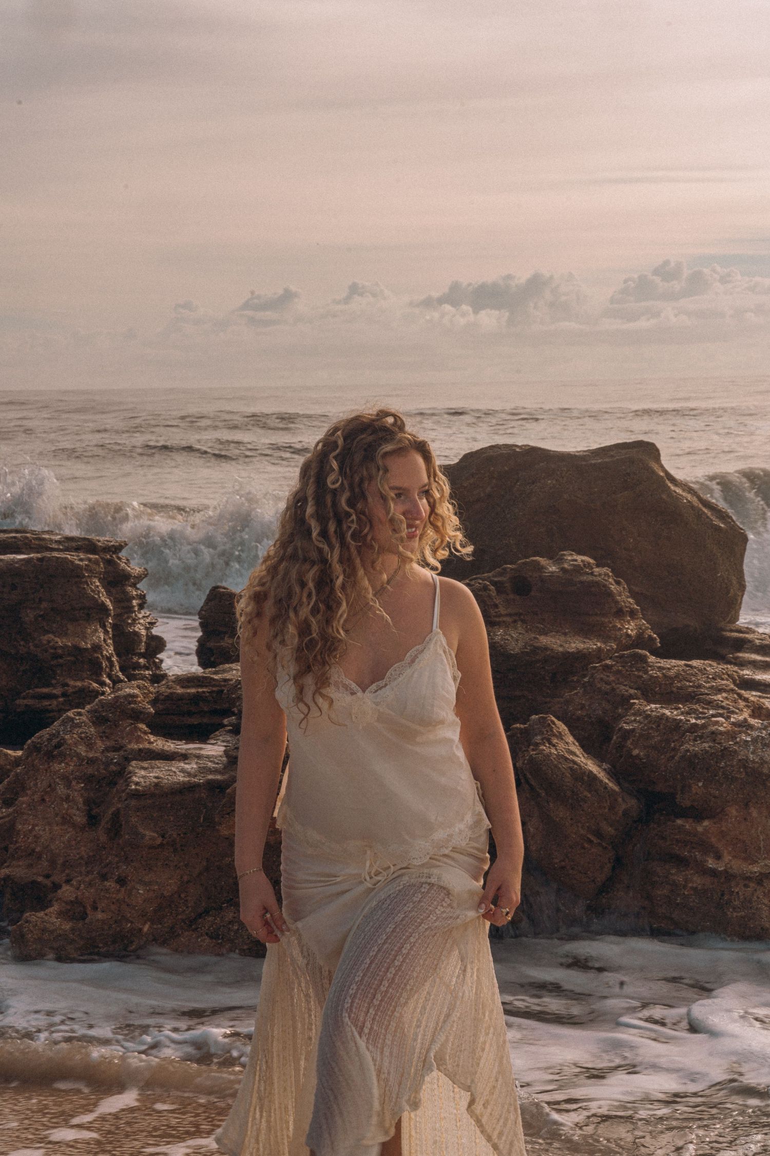 A woman in a flowing white dress stands on a rocky beach with ocean waves crashing behind her at sunset.