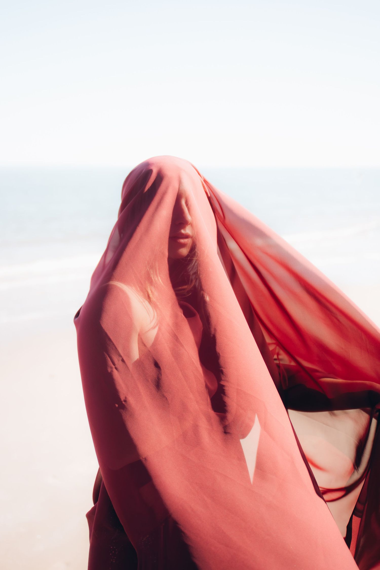 A person draped in flowing red fabric stands against a bright sky background creating an artistic silhouette effect.