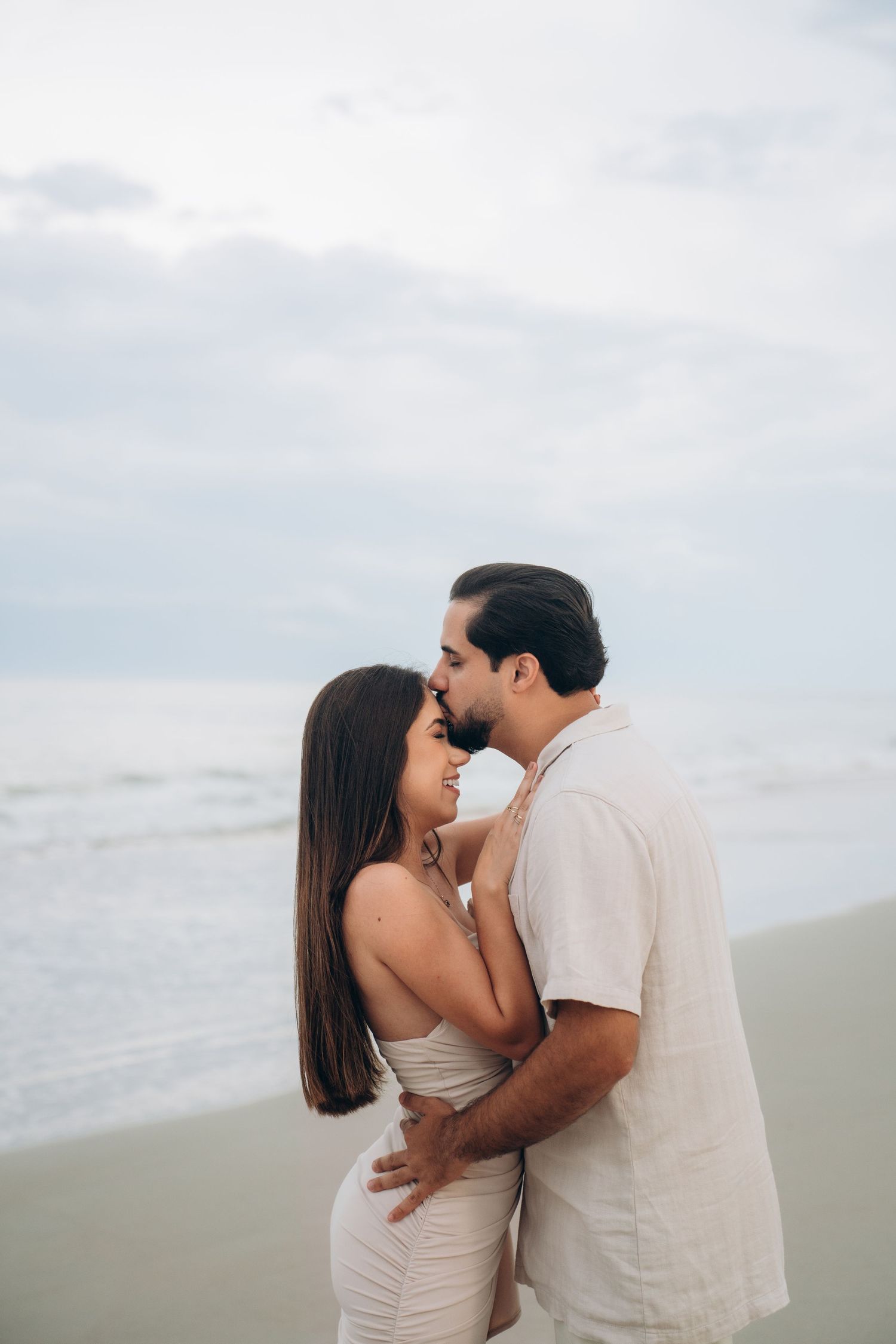 A couple in neutral colors sharing intimate moments on a cloudy beach.