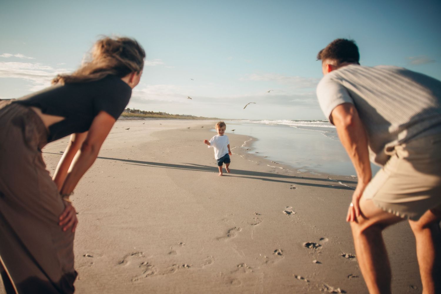 Two parents playing with a toddler on a sandy beach near the ocean shoreline on a sunny day.