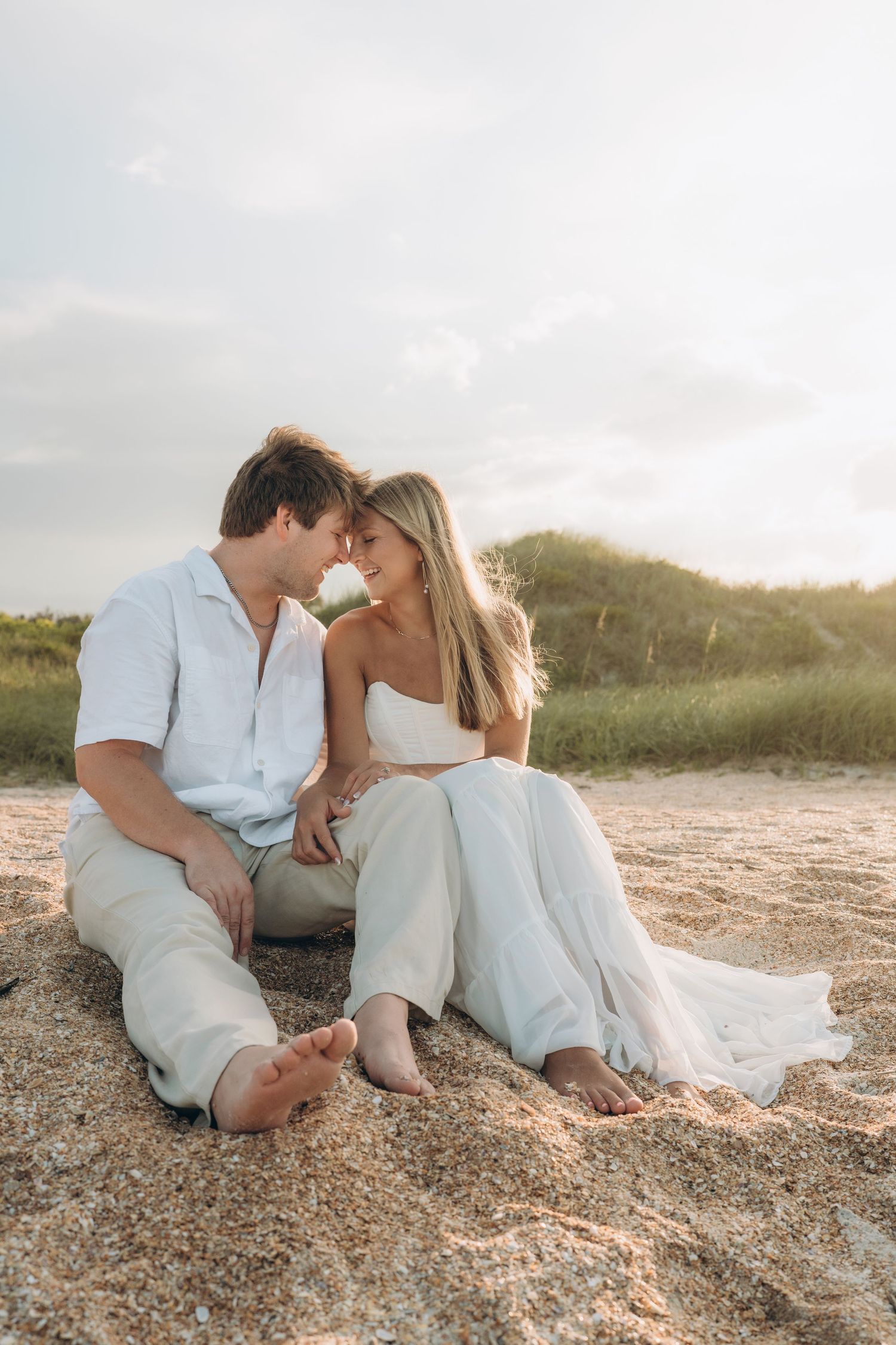A romantic couple sits together on a sandy beach at sunset wearing white and light colored clothing.