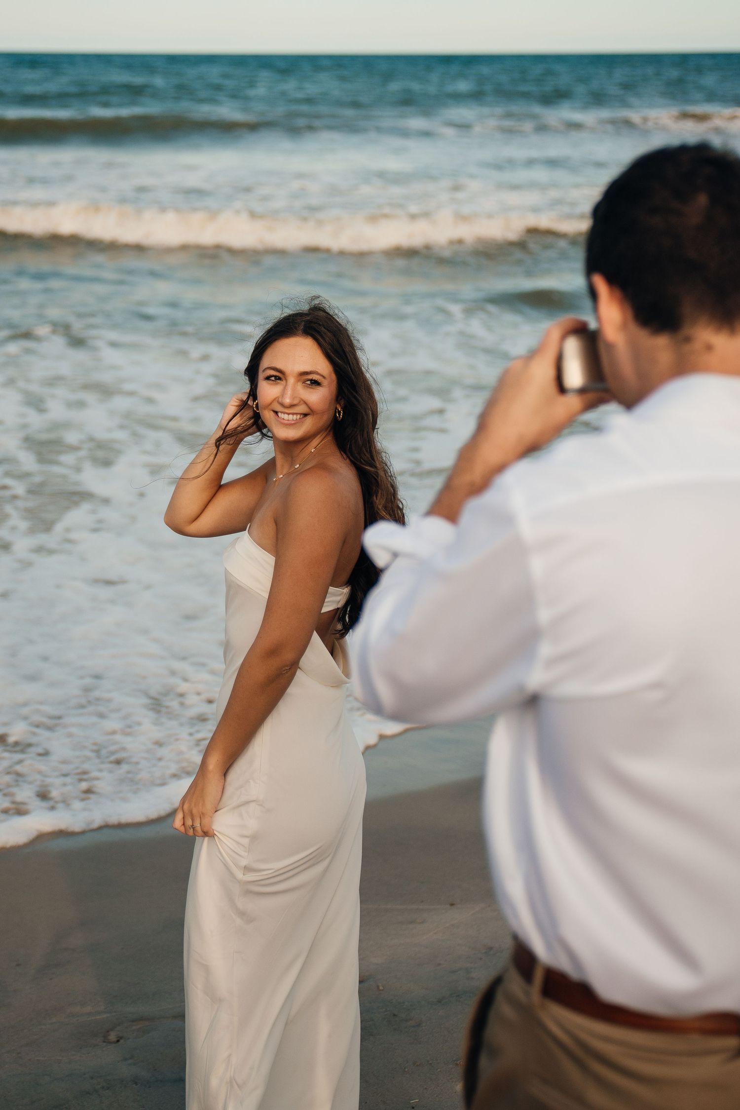 A romantic beach photoshoot with one person photographing another in a flowing white dress.