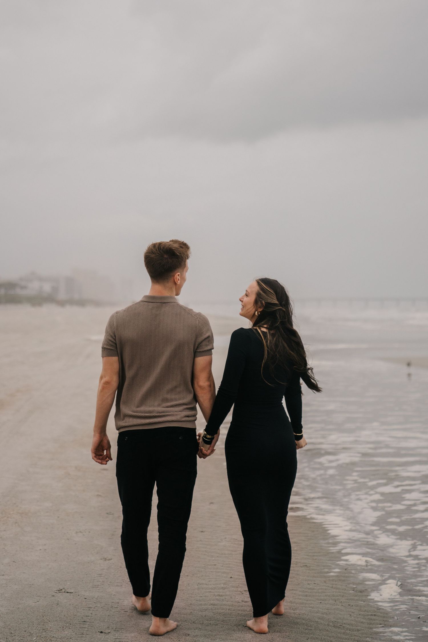 A couple walks hand in hand along the shoreline on a cloudy beach.