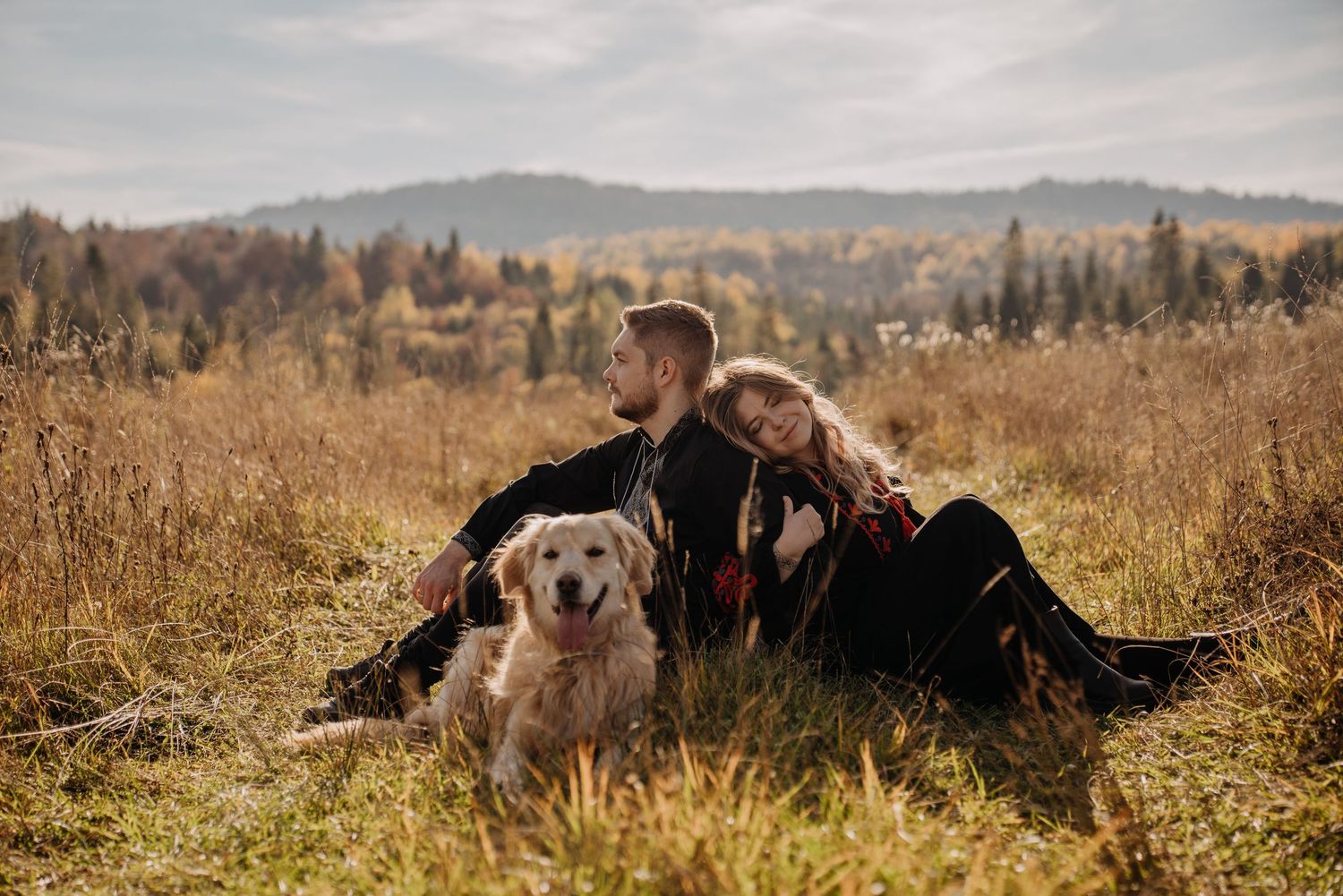 Couple sitting in autumn field with golden retriever dog, mountains and colorful fall foliage in background.