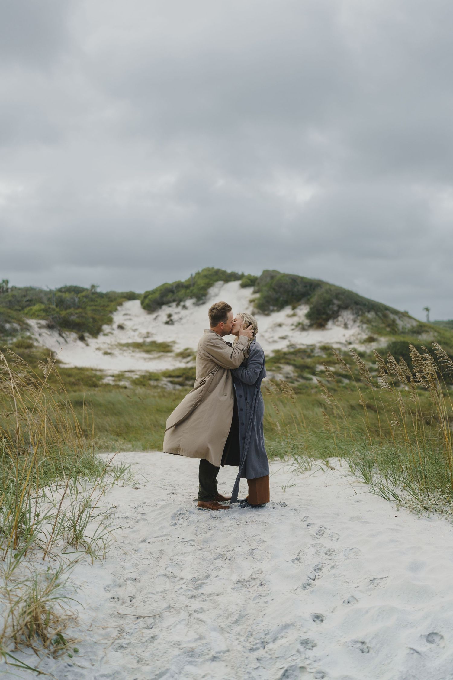 A couple embraces on a sandy beach path surrounded by sea grass and dunes on an overcast day.