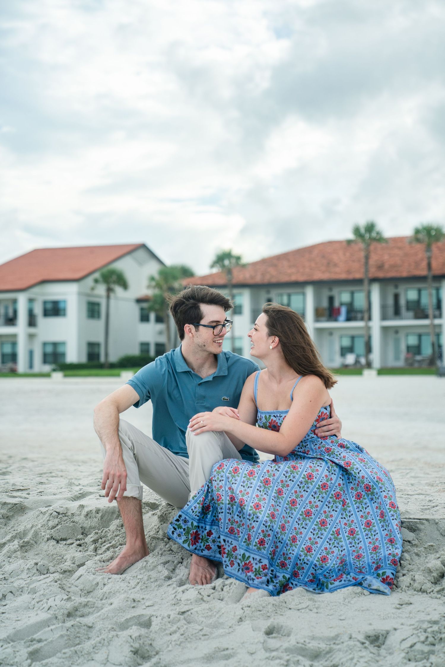 A couple sits together on a sandy beach with white coastal houses and palm trees in the background.
