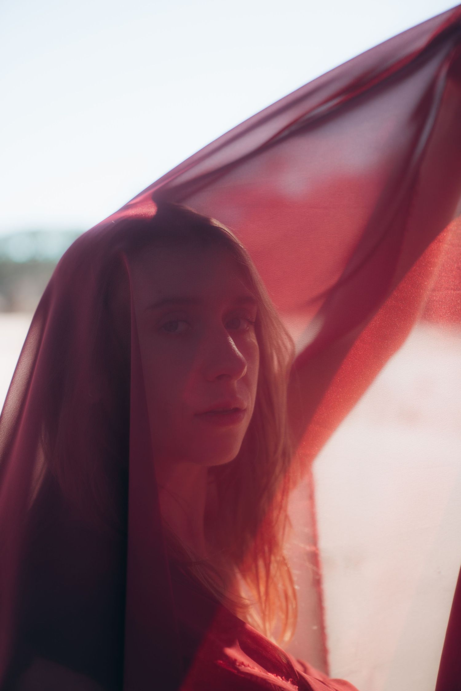 Artistic portrait through red sheer fabric creating a dreamy ethereal effect with soft lighting and shadows.