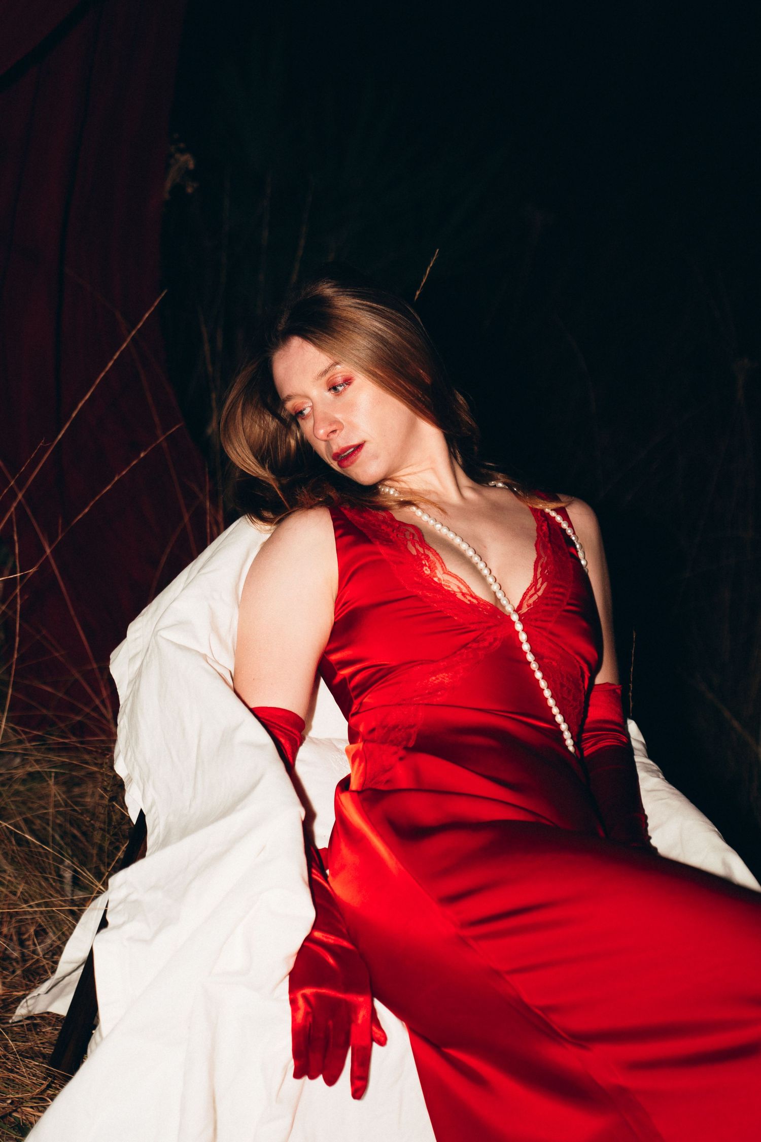 Woman in red satin gown and gloves with pearl necklace reclining outdoors at night.