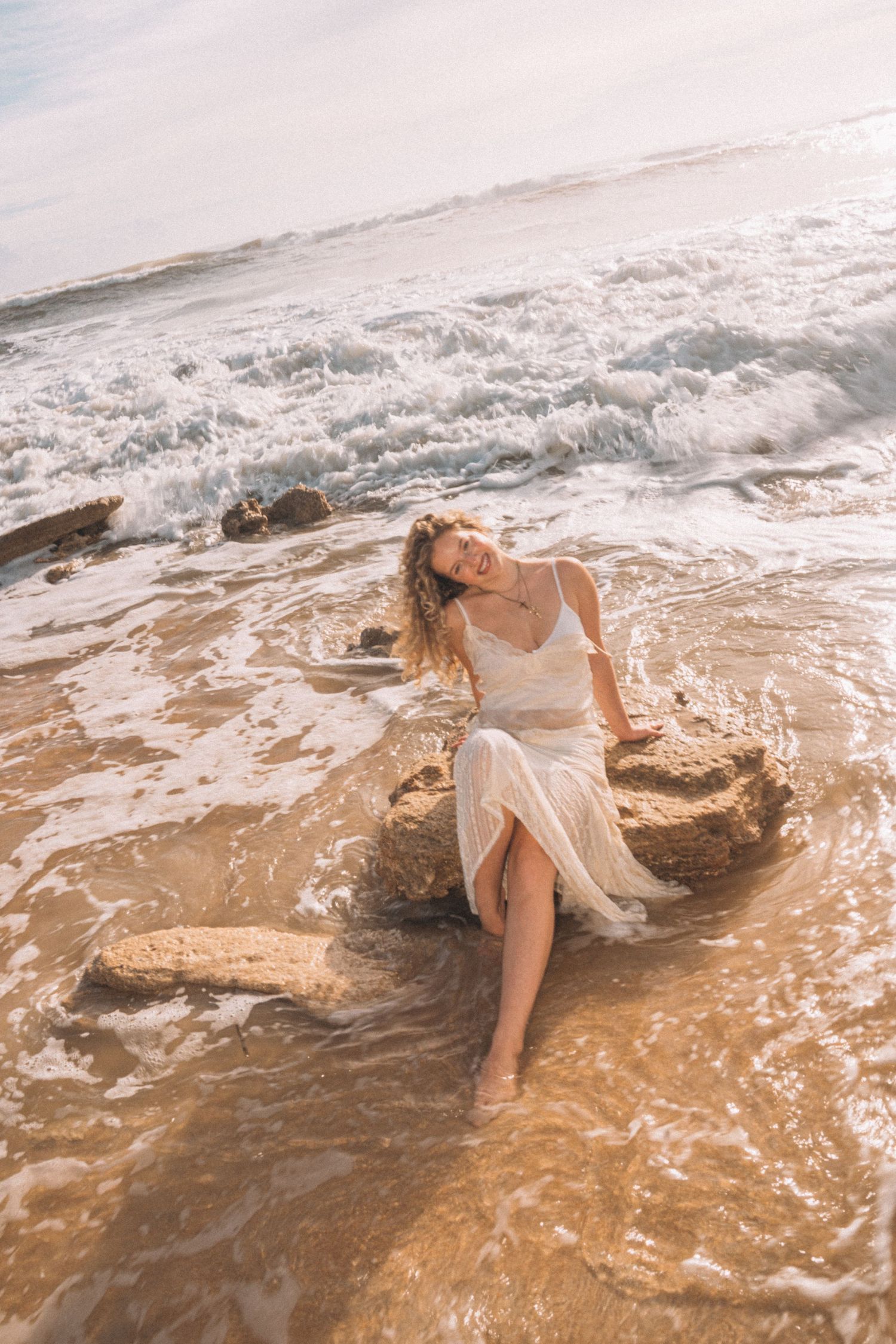 Smiling woman in white dress sitting on rock surrounded by ocean waves on sandy beach.