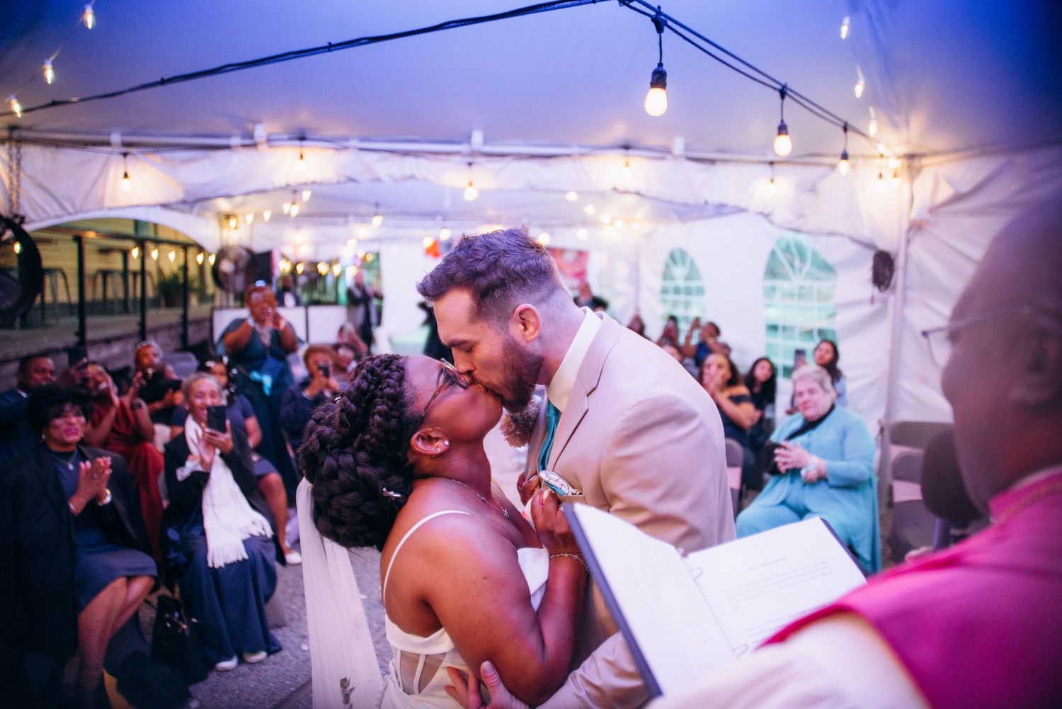 Bride and groom share first kiss under string lights in a white tent surrounded by cheering wedding guests.