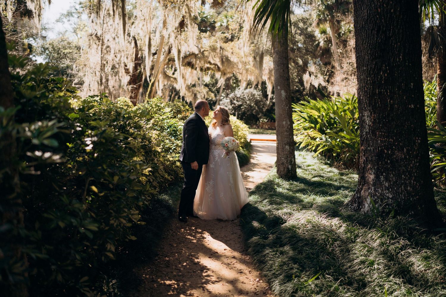 A scenic path lined with Spanish moss-draped oak trees leads through a lush Southern garden in dappled sunlight.