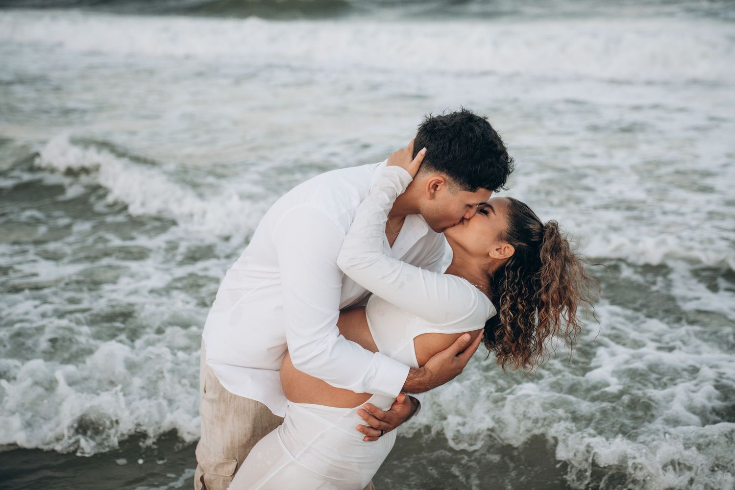 Couple sharing a romantic kiss on the beach, dressed in white, with ocean waves crashing around them.