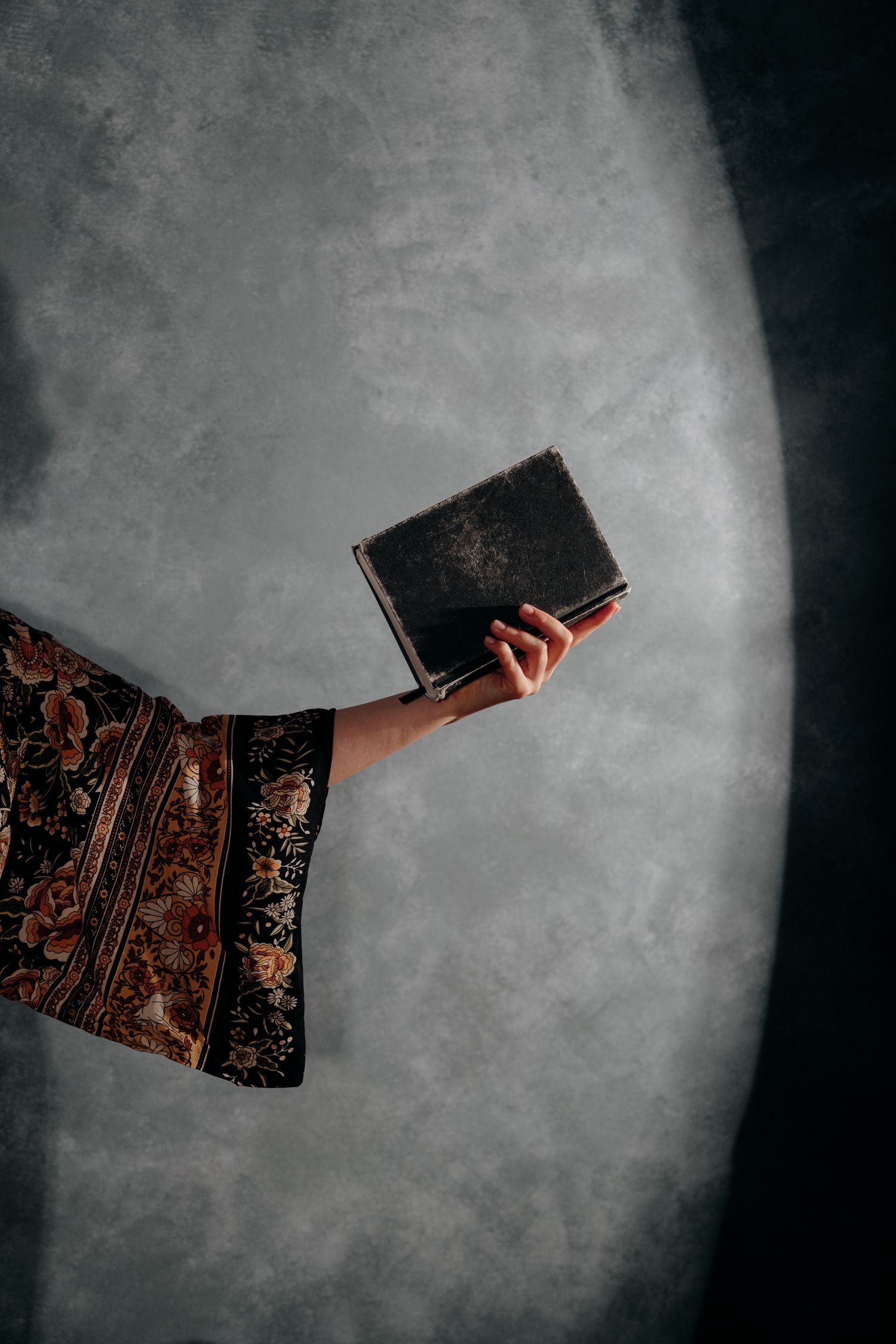 A hand in a floral robe holds an old dark book against a moody gray background.
