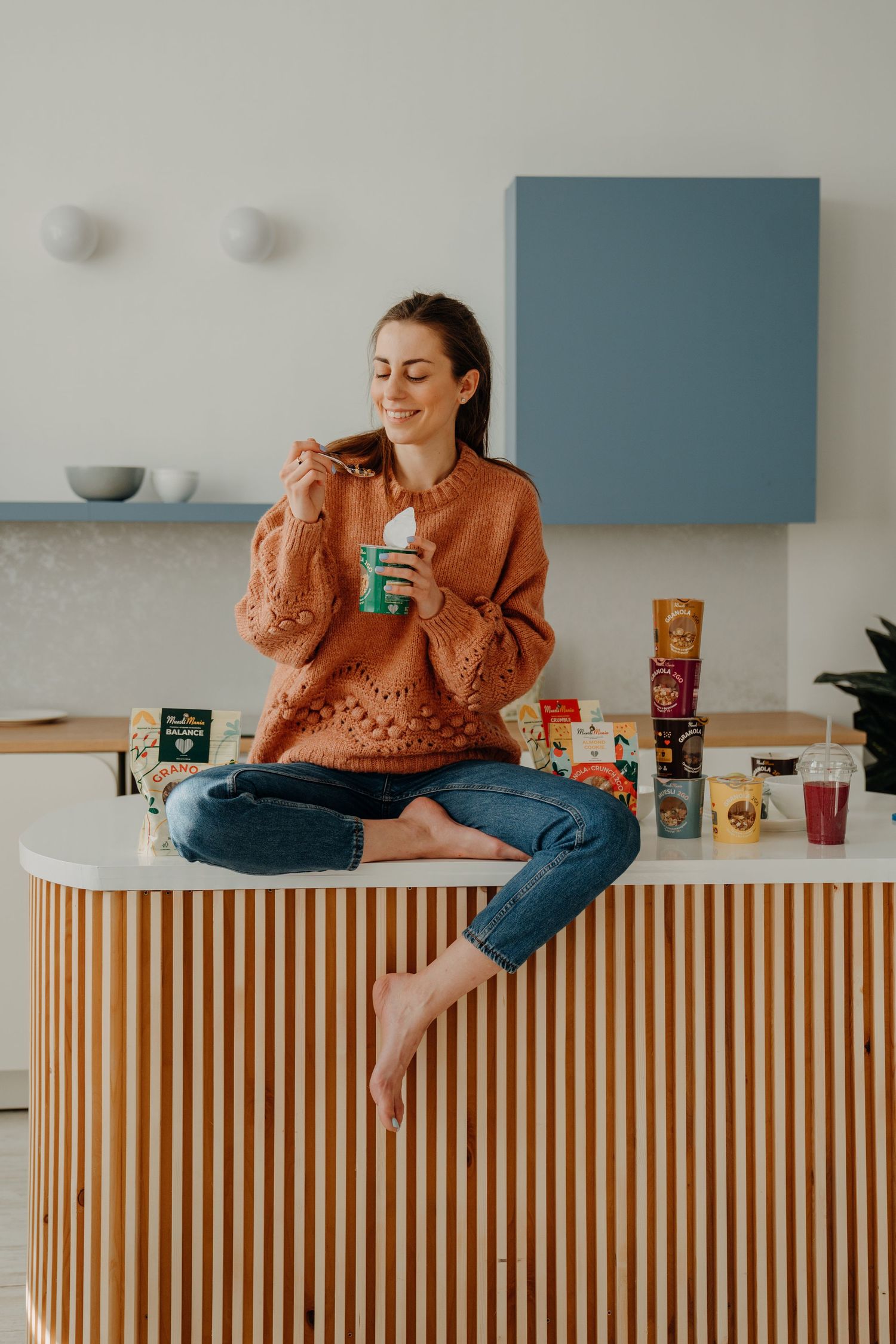 Smiling woman in orange sweater sitting on kitchen counter eating yogurt surrounded by healthy food products.