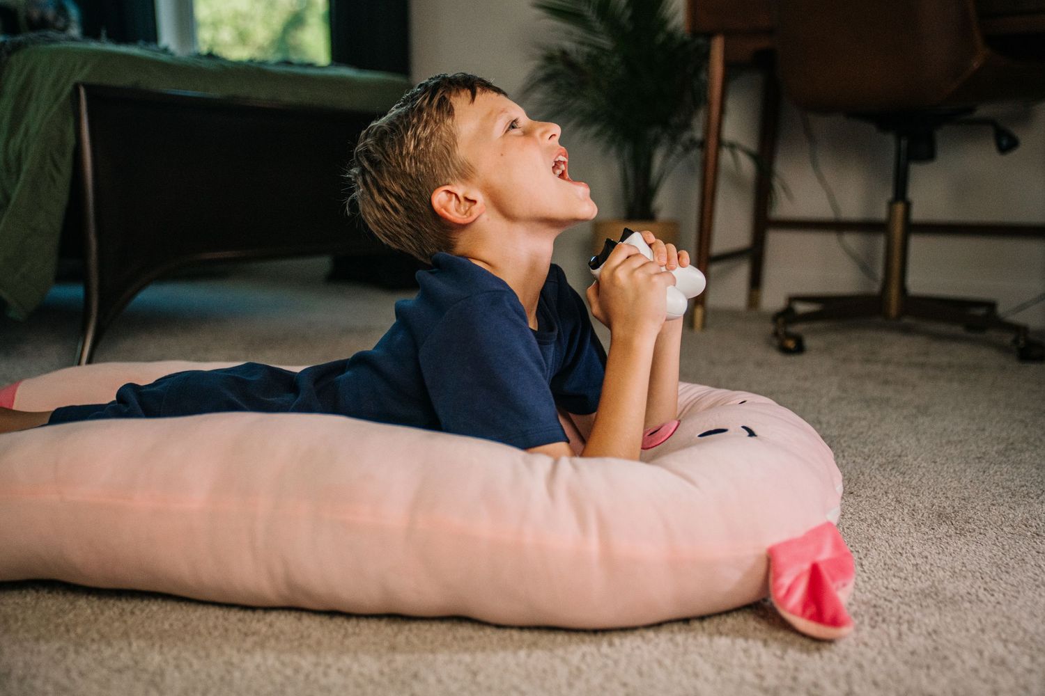 Boy lying on a large pink stuffed animal pillow on carpet, laughing with joy indoors.