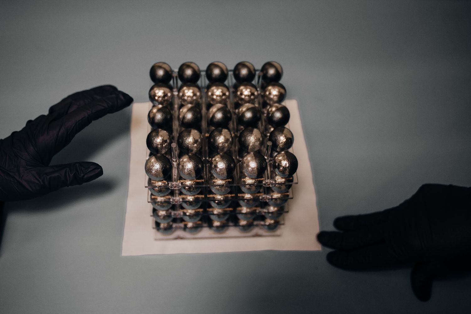 Hands in black gloves holding a cube made of metallic magnetic balls arranged in a grid pattern on a white surface.