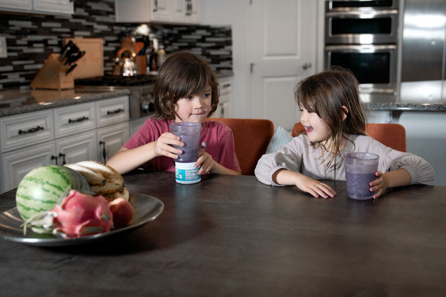 Two children sitting at a kitchen table sharing drinks with a fruit bowl nearby.
