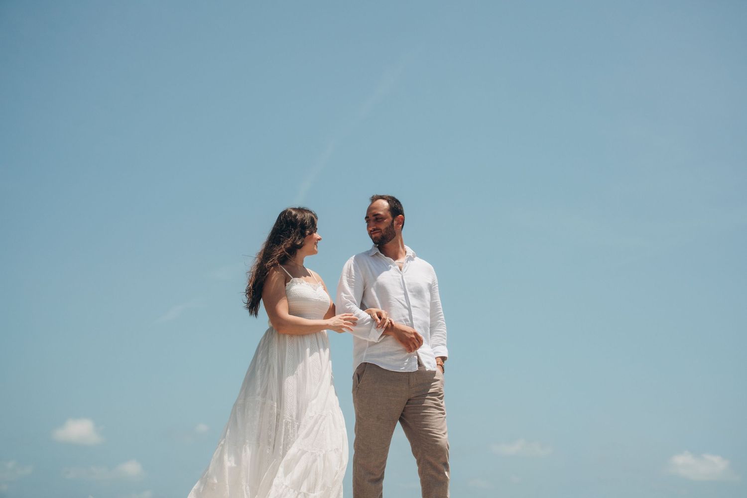 Bride in white dress and groom in white shirt hold hands under bright blue sky outdoors.