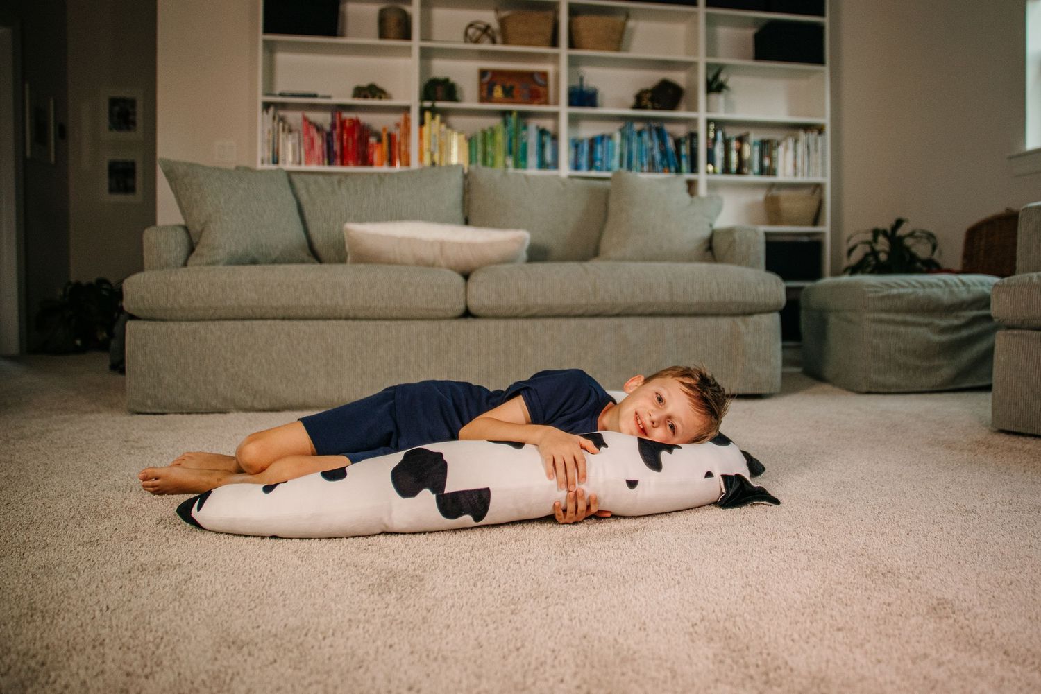 Child lying on carpet hugging a large black and white cow-print body pillow in a cozy living room.