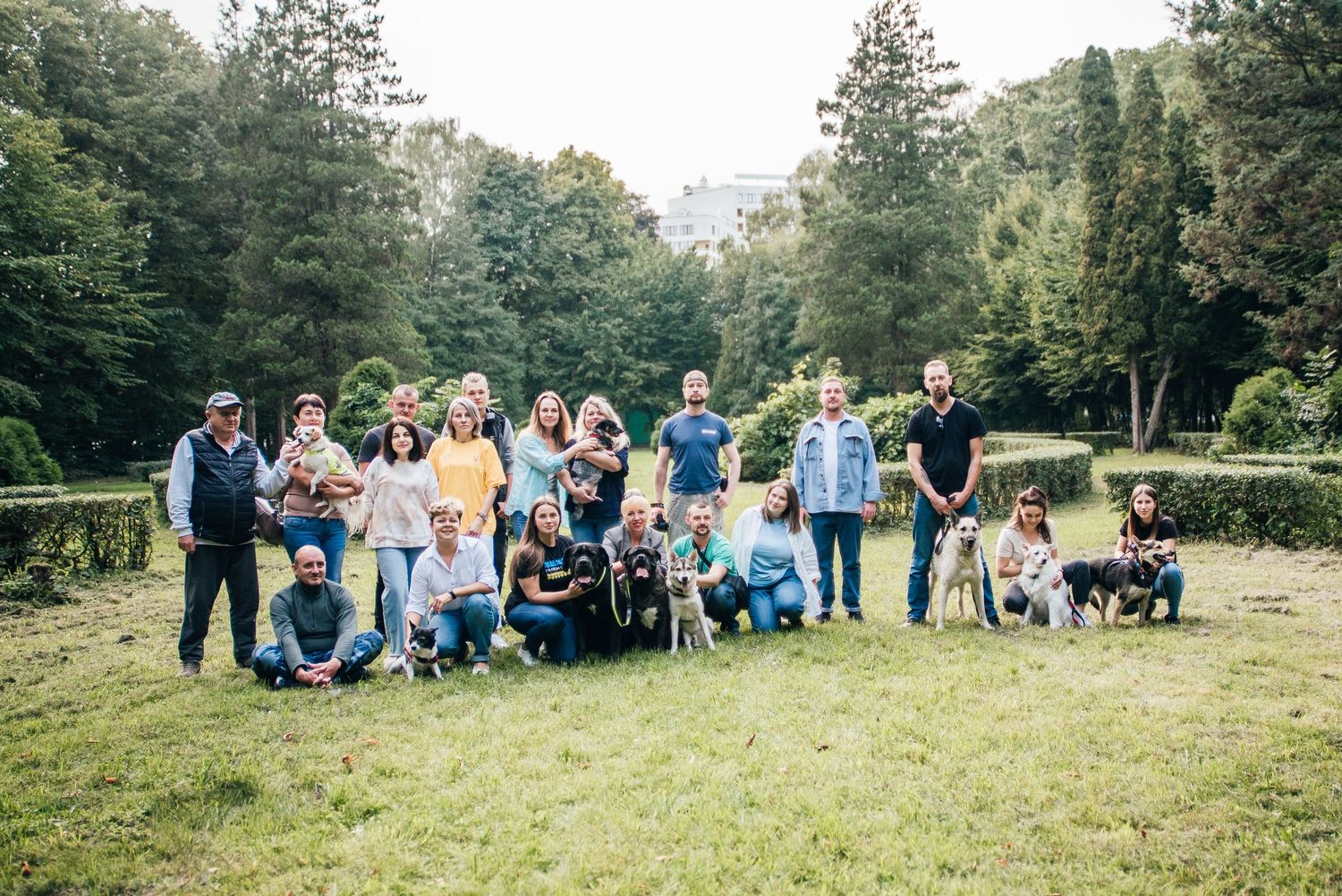 Large group of people with their dogs posing together on a green lawn surrounded by trees in a park.