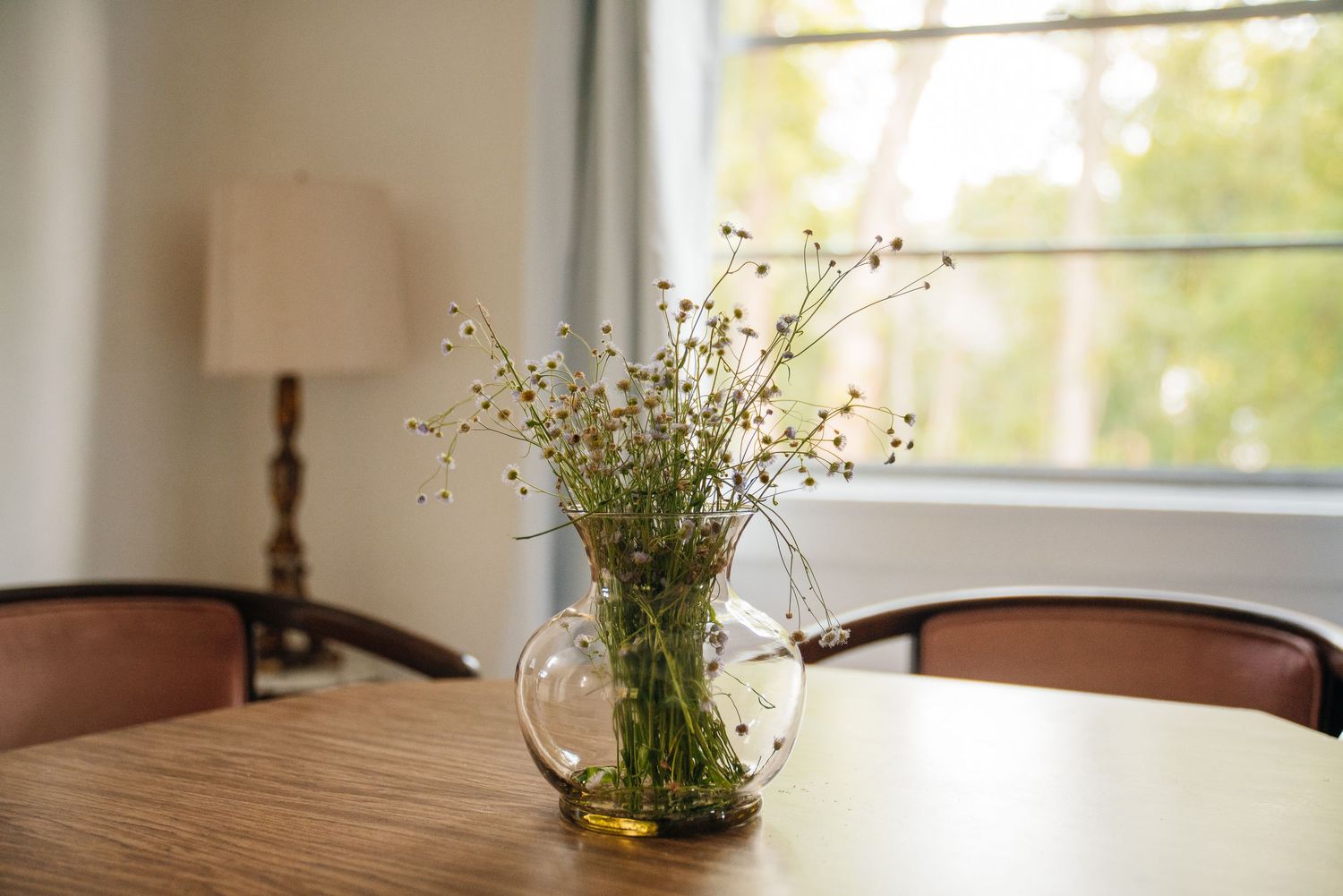 Glass vase with wildflowers on wooden dining table near bright window with lamp.