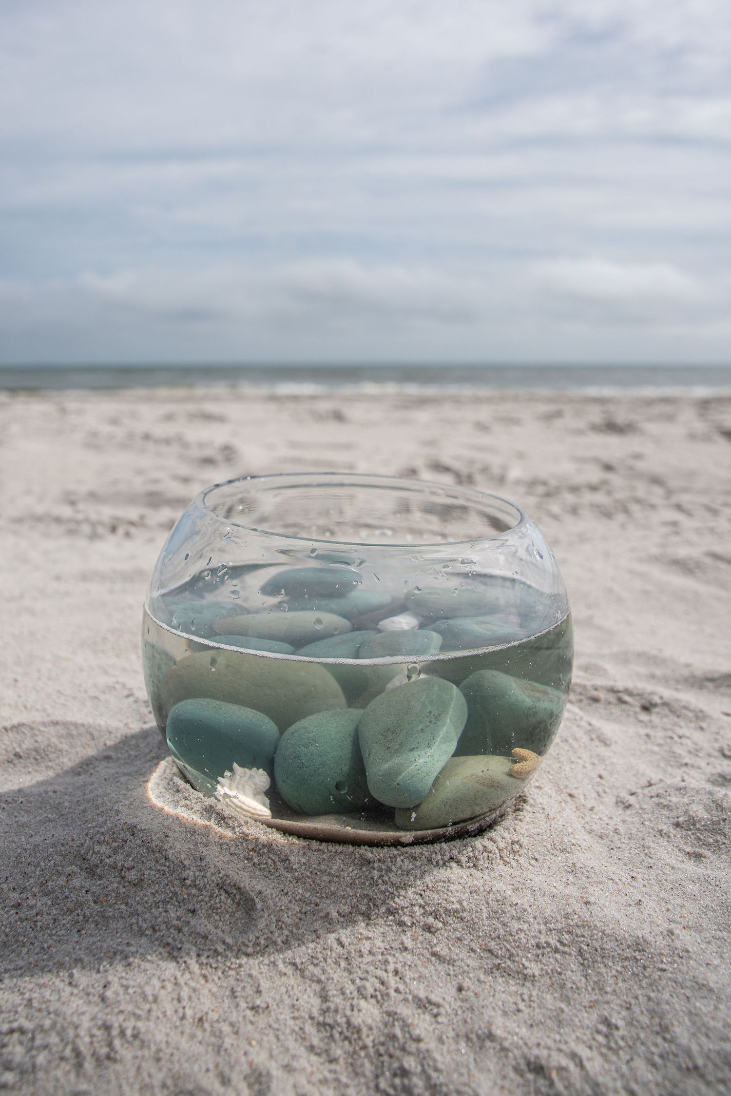 Glass bowl filled with green stones and water sitting on sandy beach with ocean horizon in background.