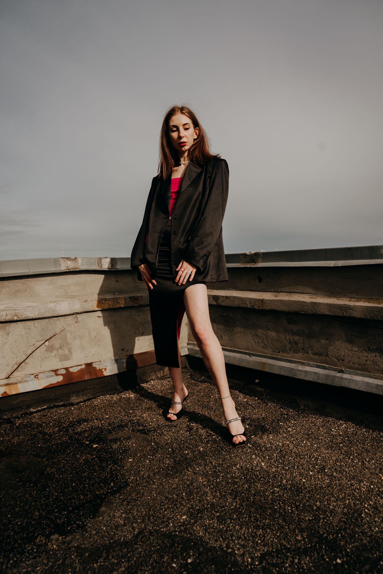 Young woman in black blazer and skirt poses on rooftop wearing heels.