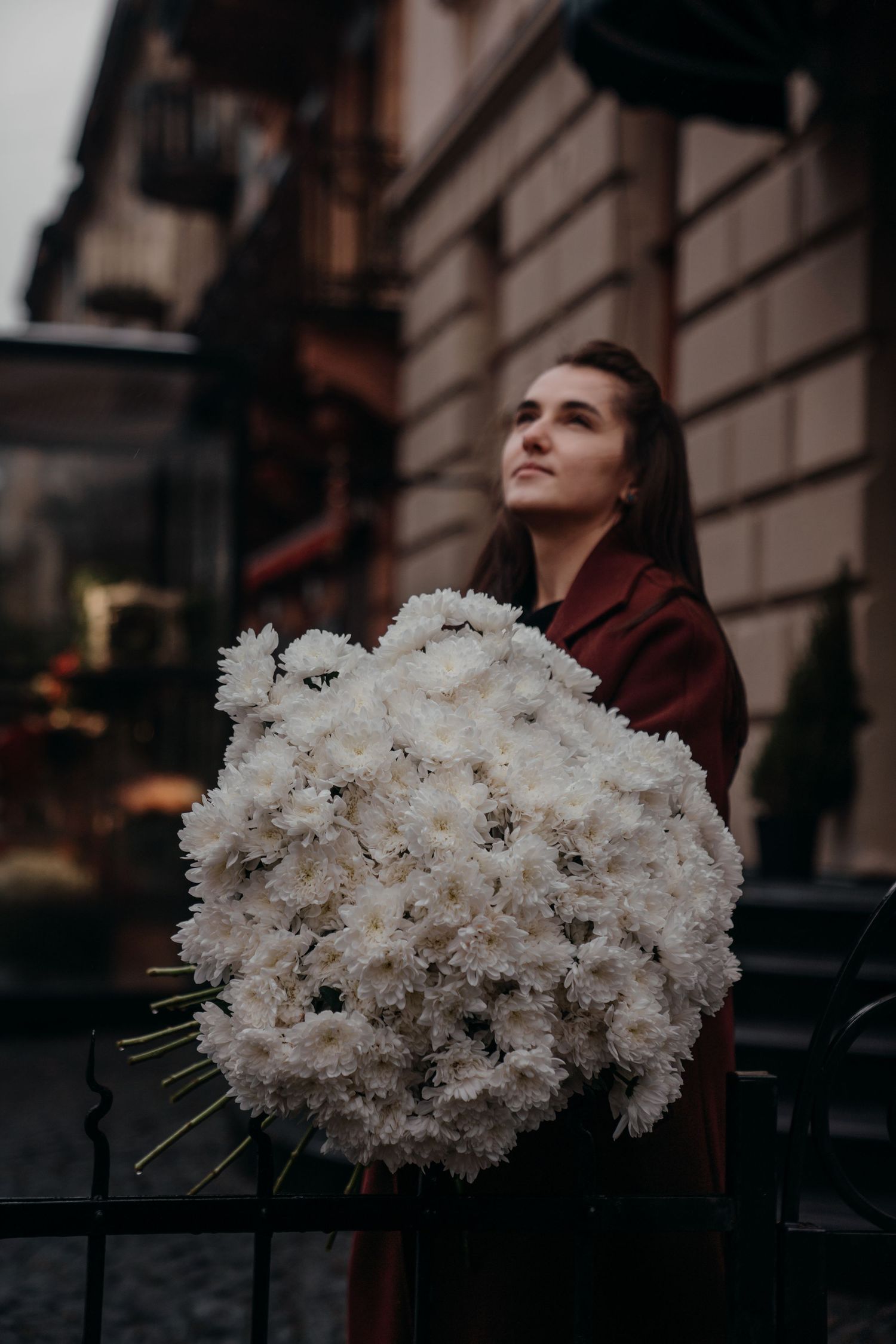 Woman in dark red coat holding oversized bouquet of white chrysanthemums on urban street.