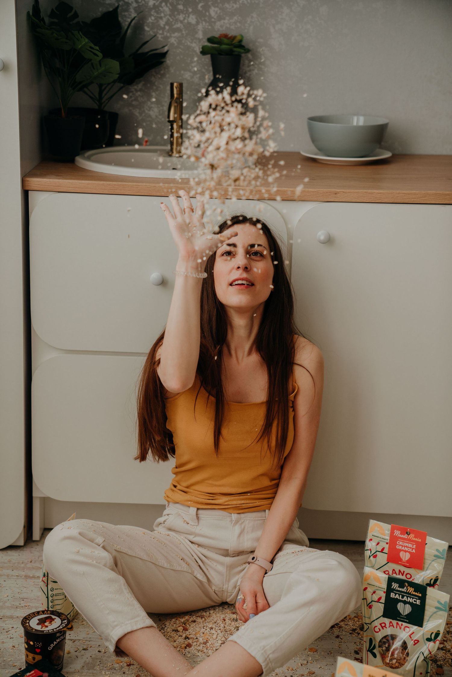 A young woman sitting on a kitchen floor tossing granola in the air playfully.