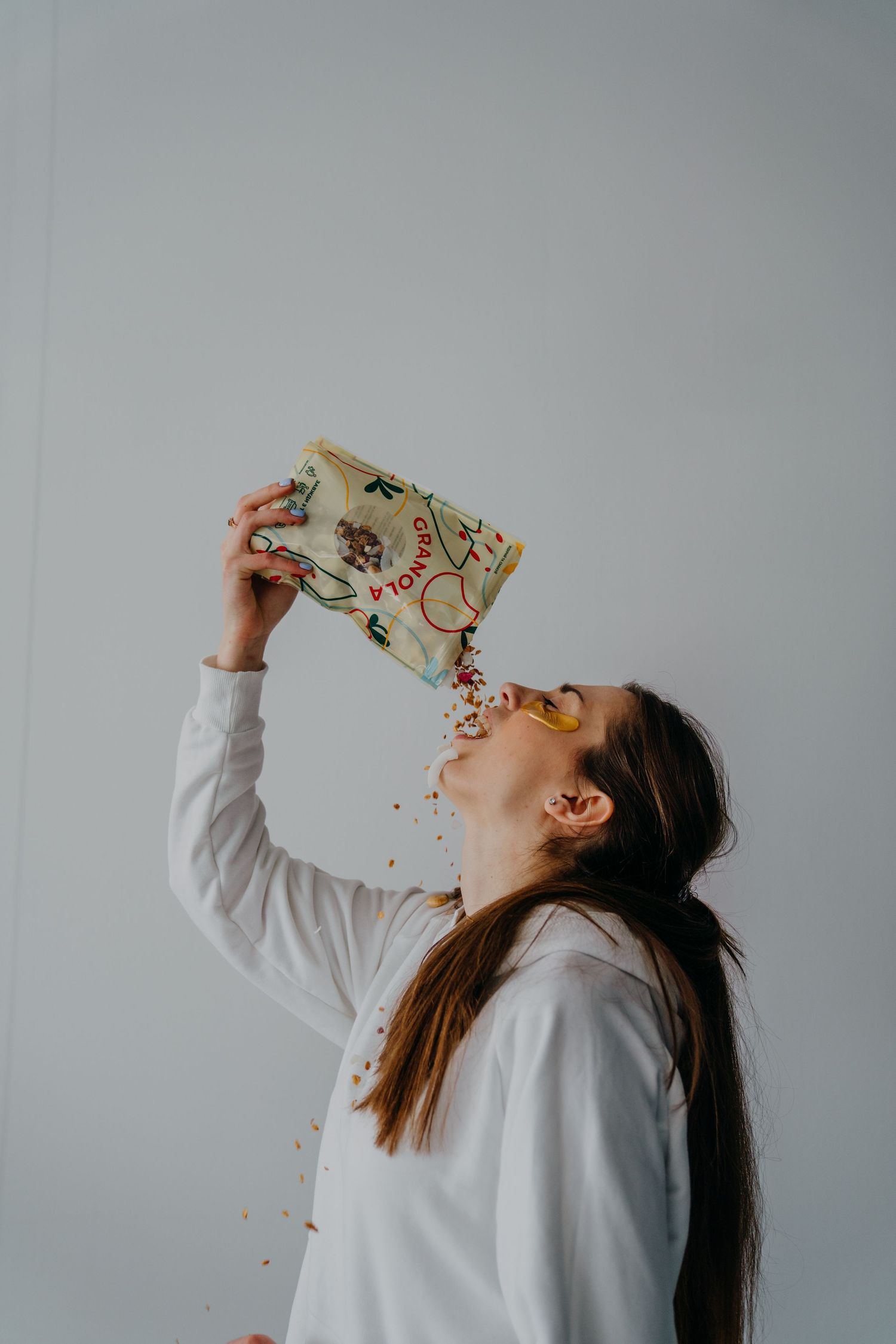 A young woman pouring granola directly from a colorful bag into her mouth against a white background.