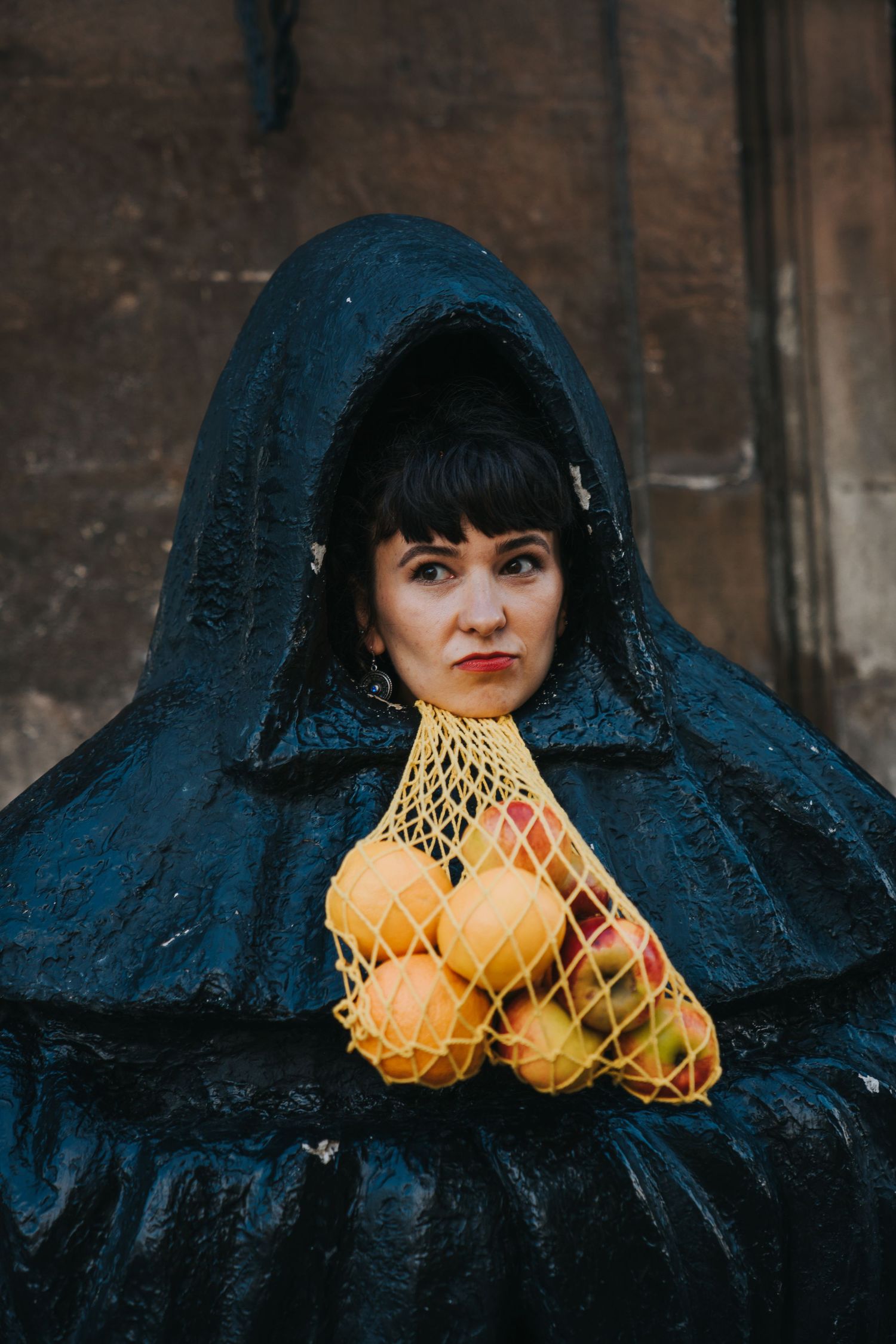 Woman in black hooded cloak holding a mesh bag of fresh oranges and apples against a stone wall.