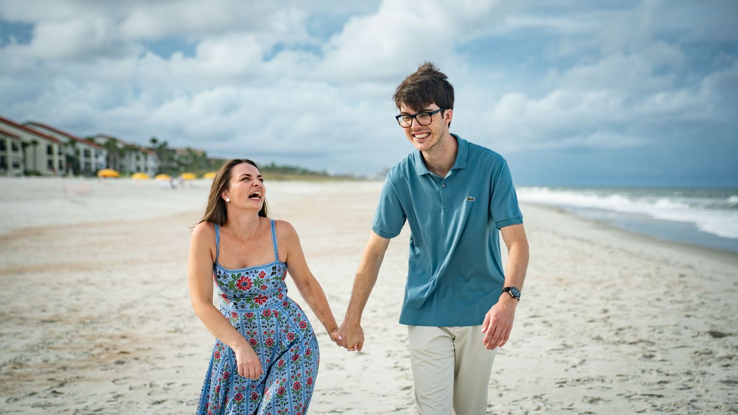 Couple walking hand in hand along a scenic beach shoreline with waves and blue sky in the background.
