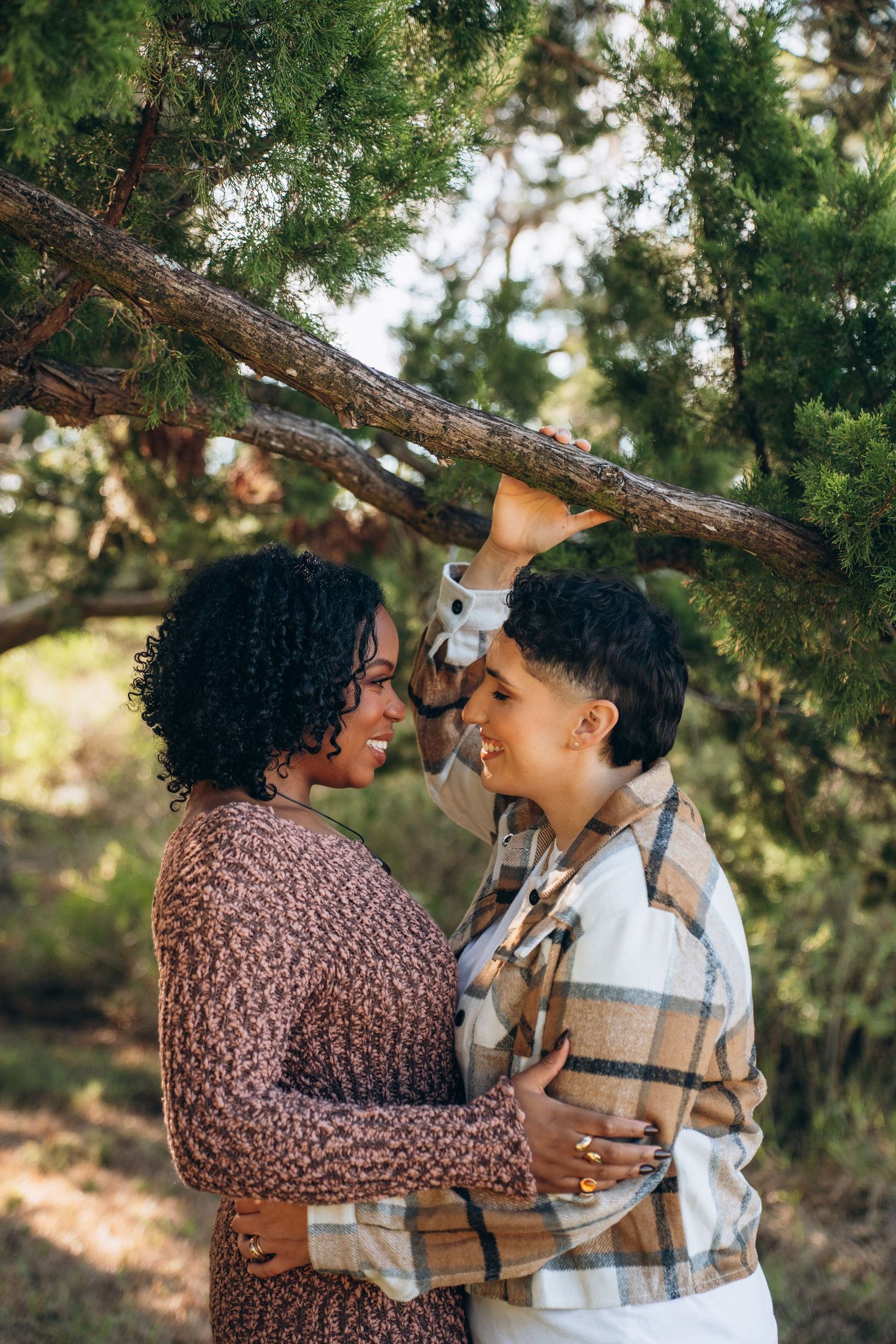 A couple shares an intimate moment outdoors under evergreen trees on a sunny day.