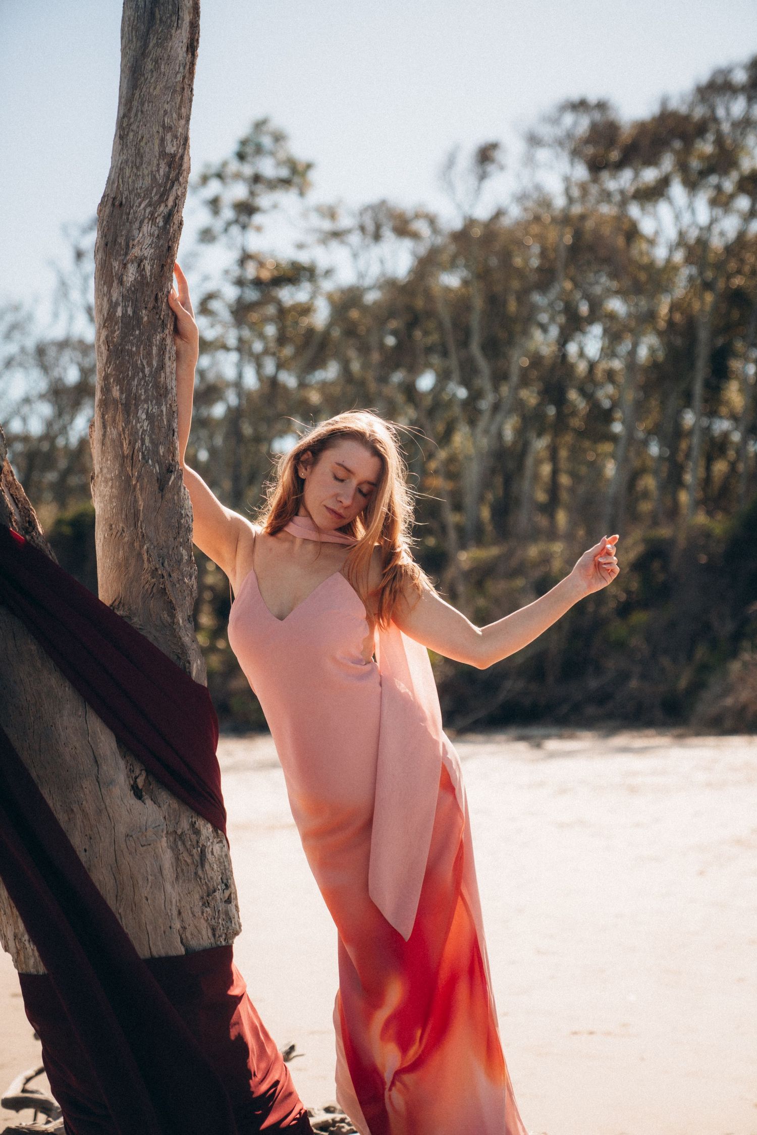 Flowing coral dress catches sunlight near weathered tree trunk on sandy beach.