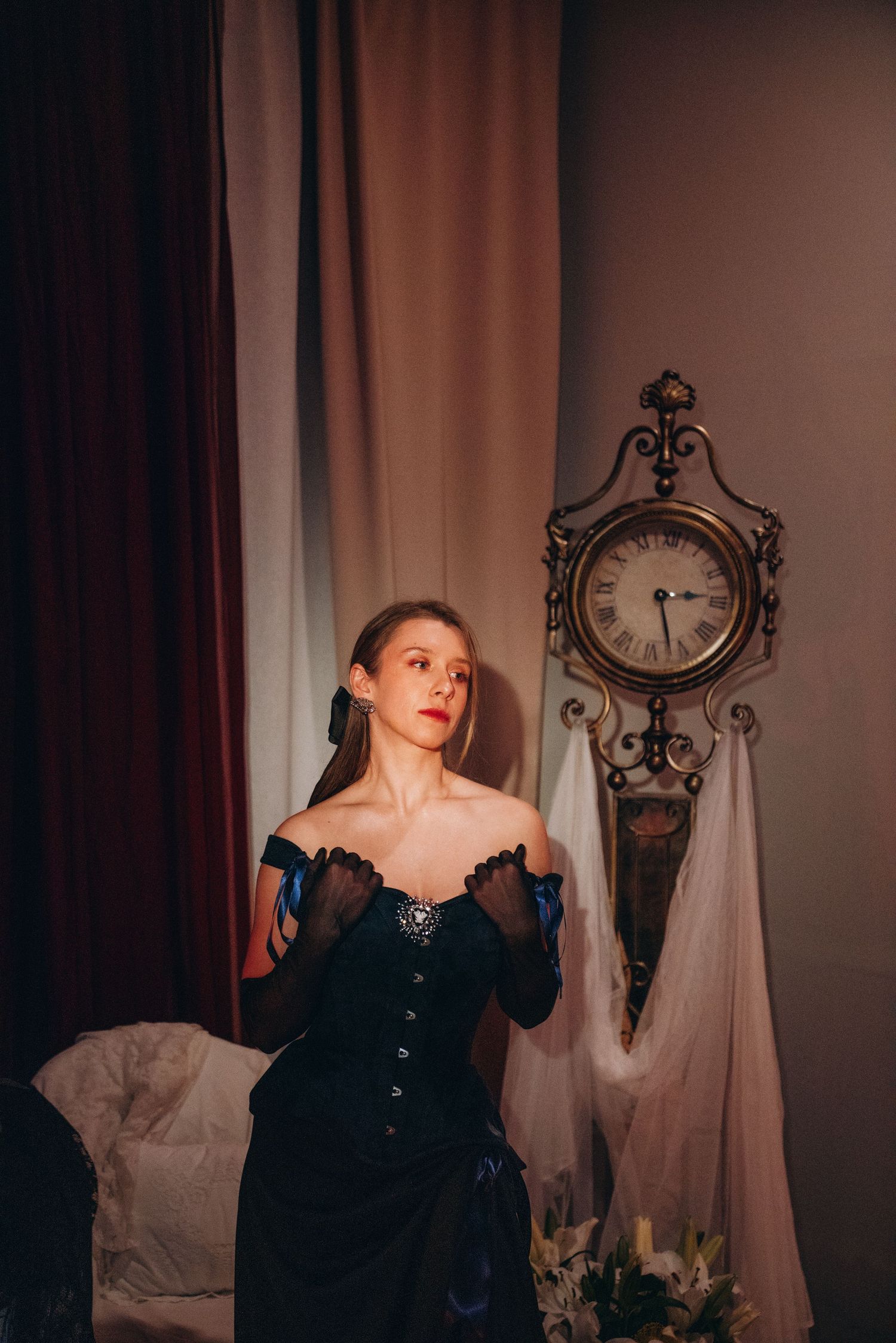 Young woman in black Victorian corset dress with gloves poses beside ornate antique clock in dramatic lit room.