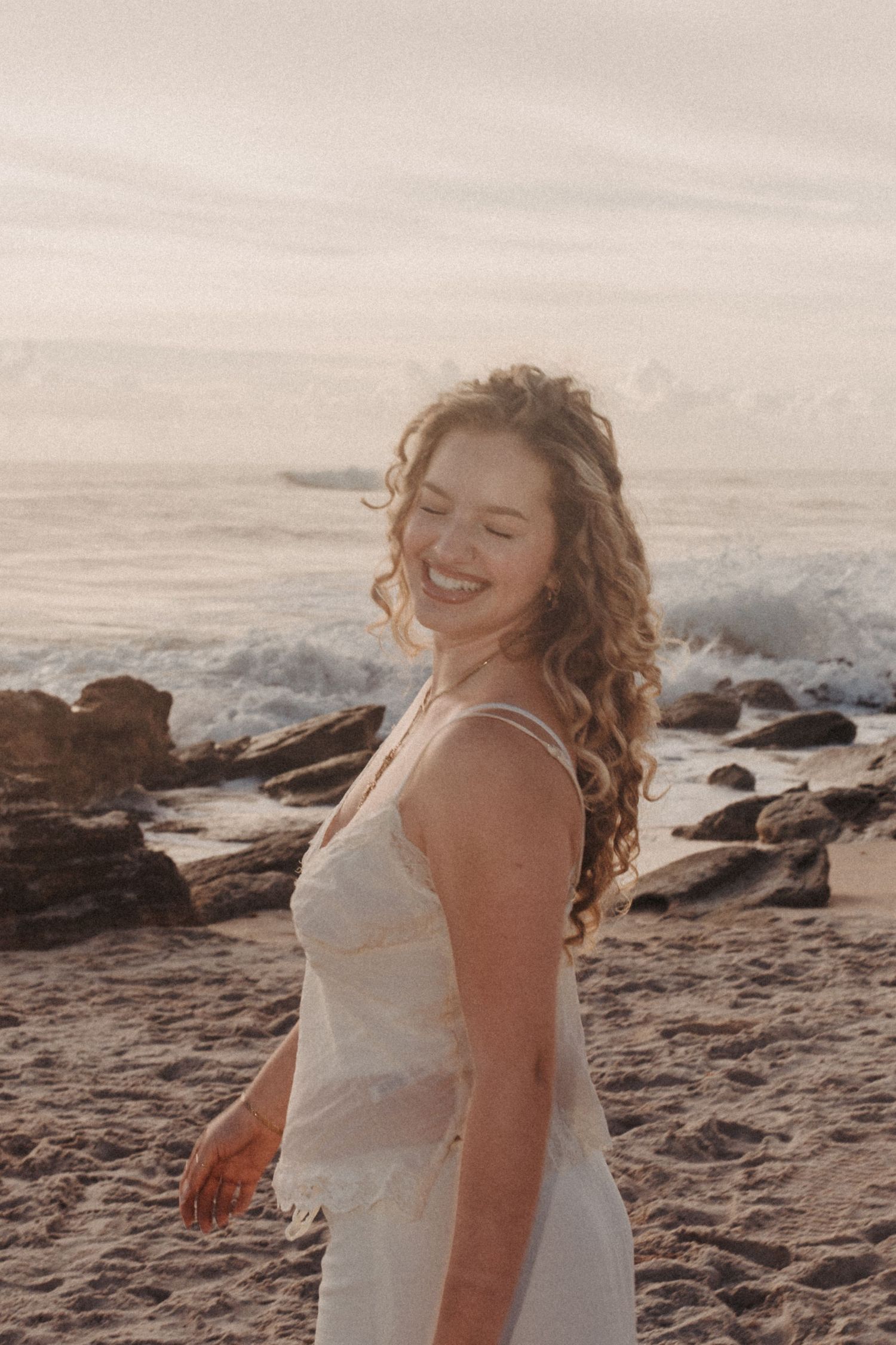 Smiling young woman with curly hair in a white dress standing on a sandy beach with rocky shores and ocean waves.