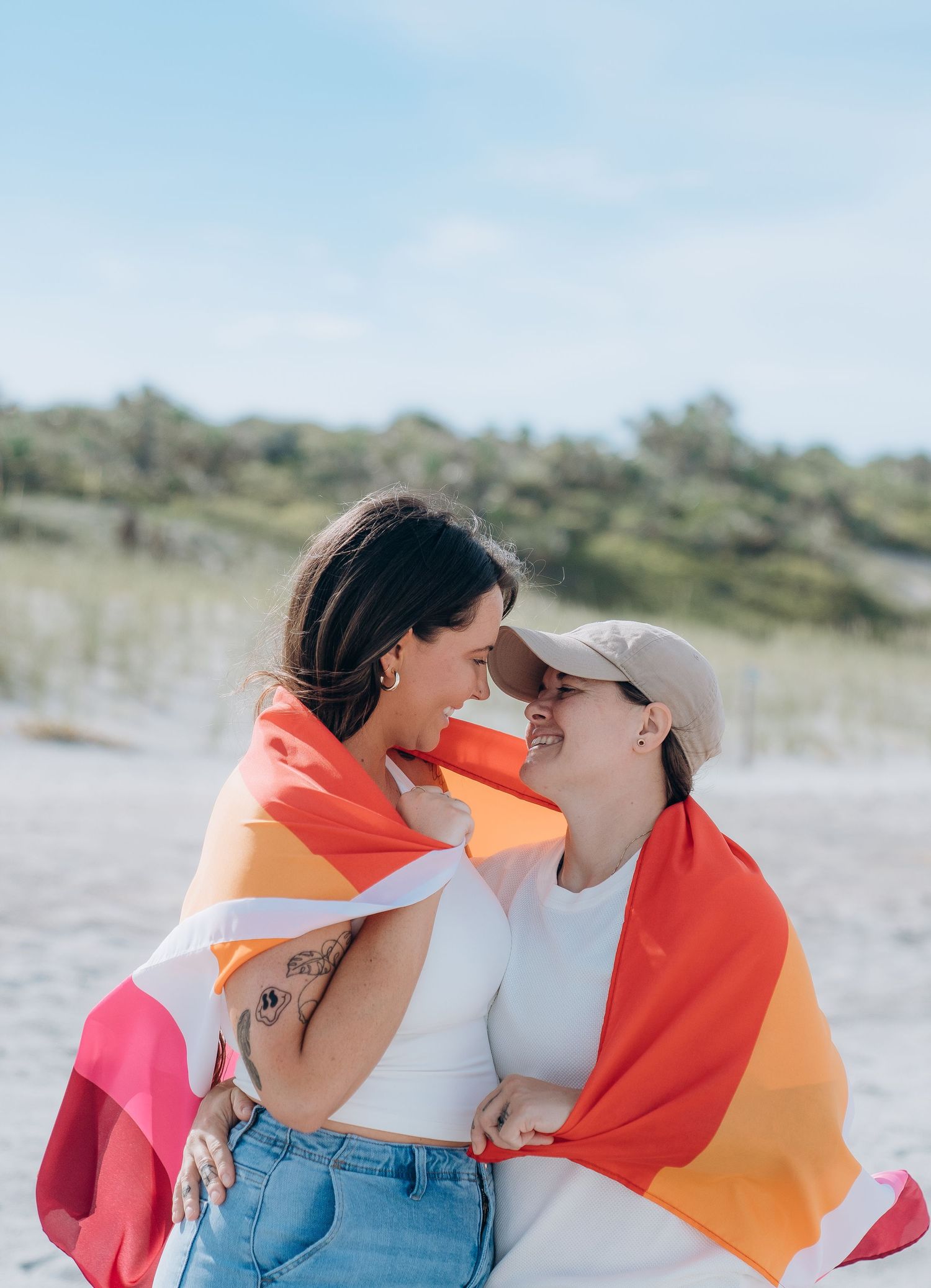 A couple draped in a colorful pride flag embracing on a sunny beach.