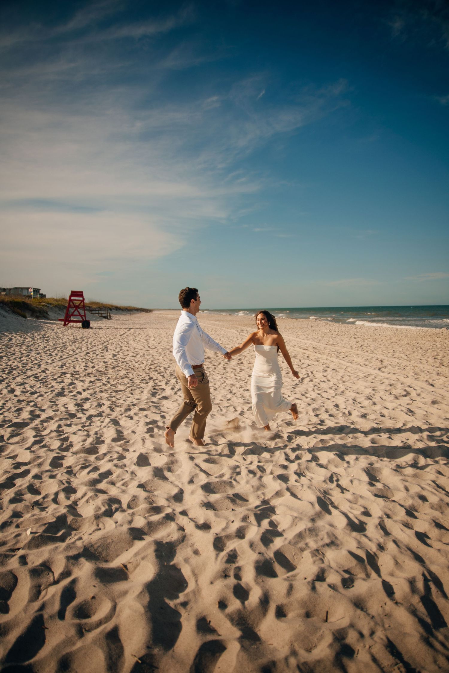 Playful couple runs and dances together on a sunny beach with footprints in the sand.
