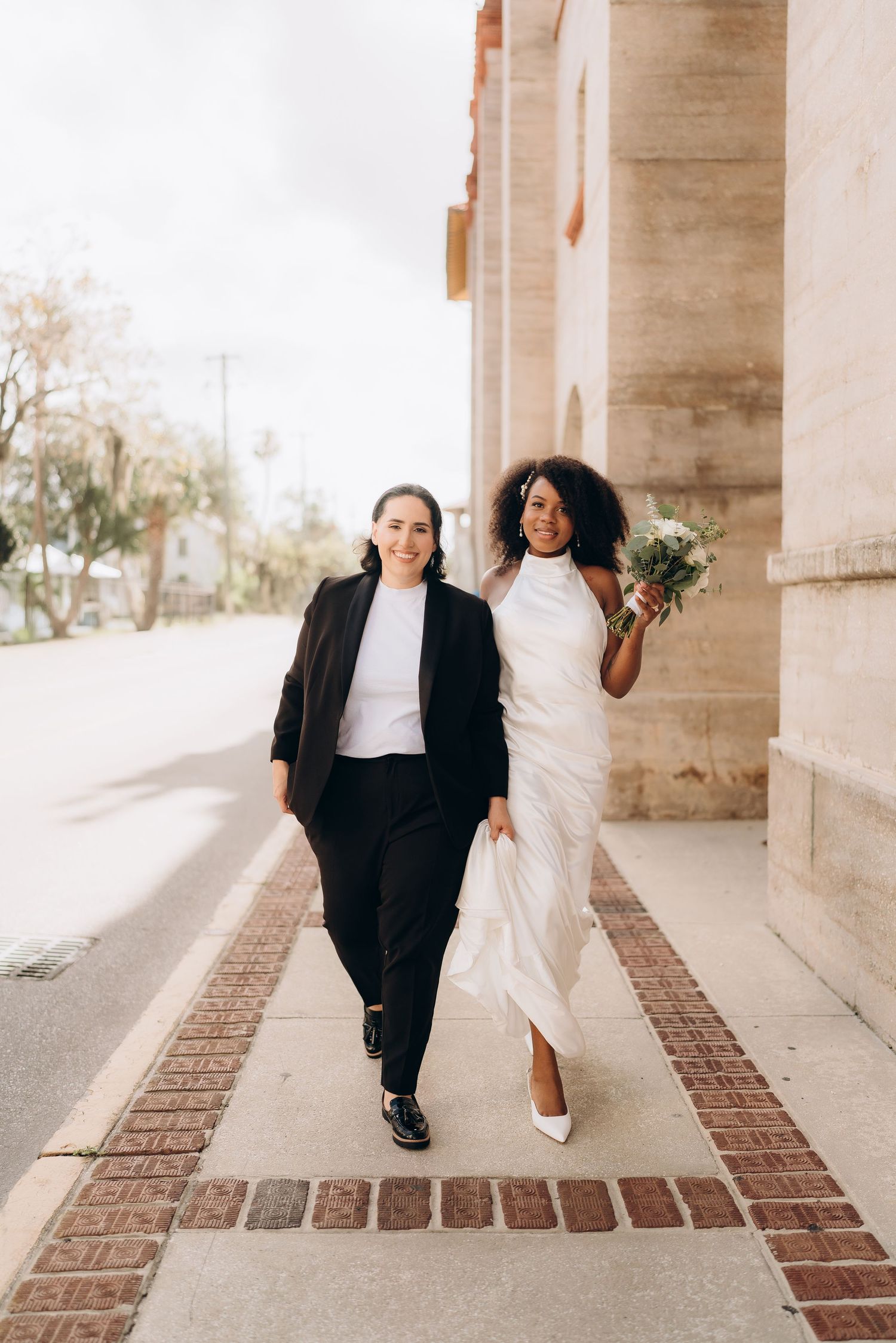 A couple in formal attire walks playfully along a brick-paved walkway.