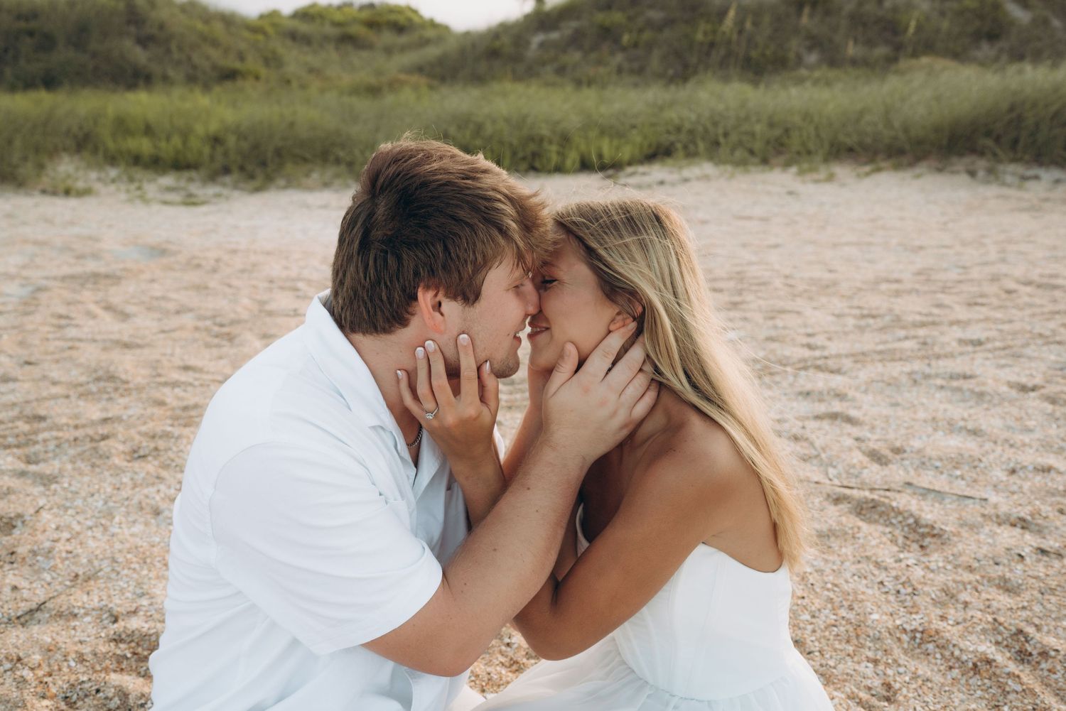 A couple shares an intimate moment on a sandy beach with dunes in the background.