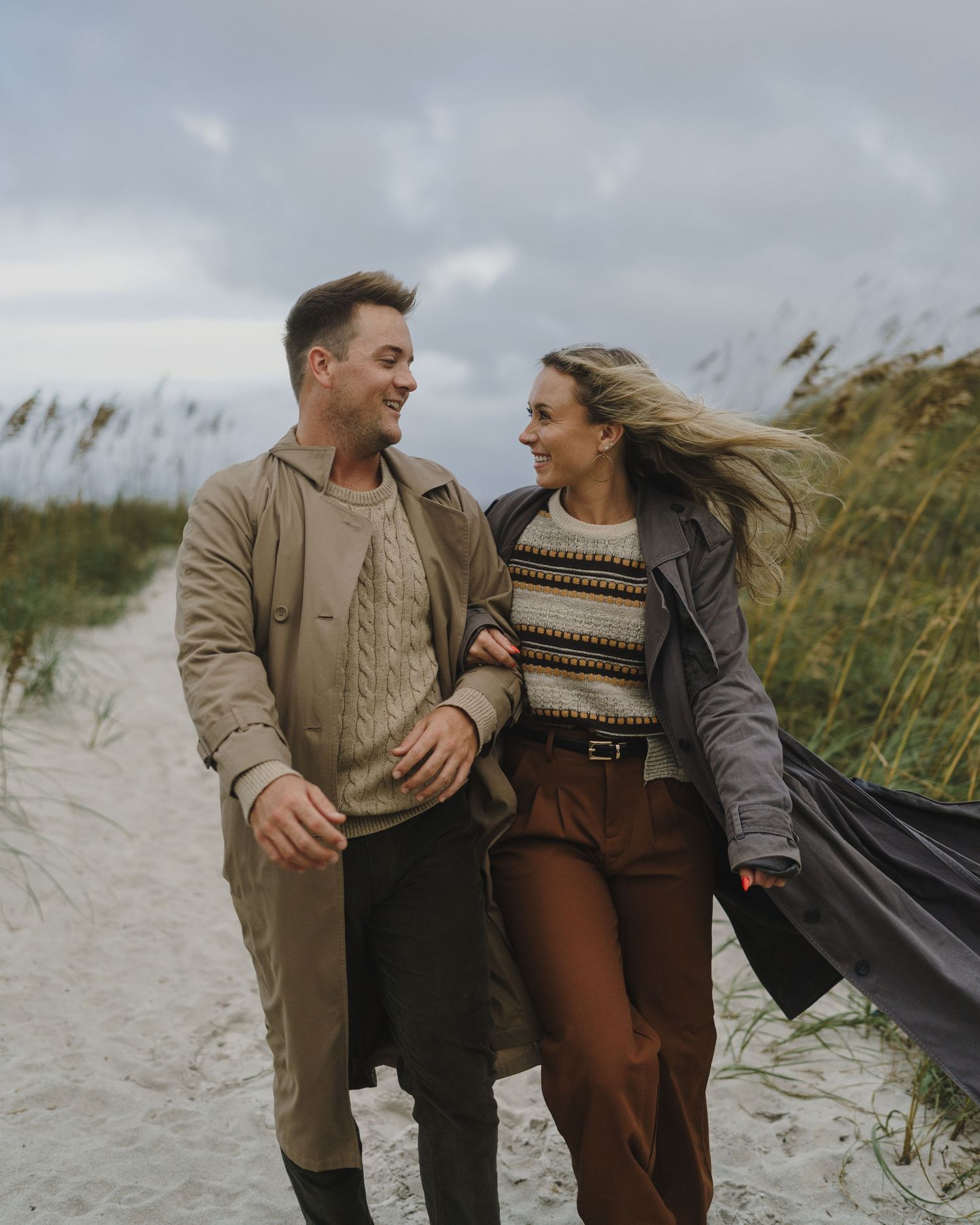 Couple walking together on windy beach path wearing casual fall outfits.