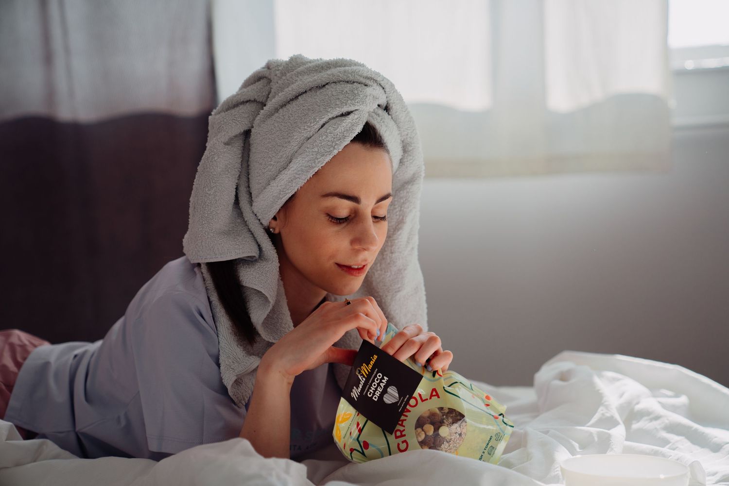 Woman in towel turban and robe lying on bed reading a snack package in a bright, cozy bedroom.