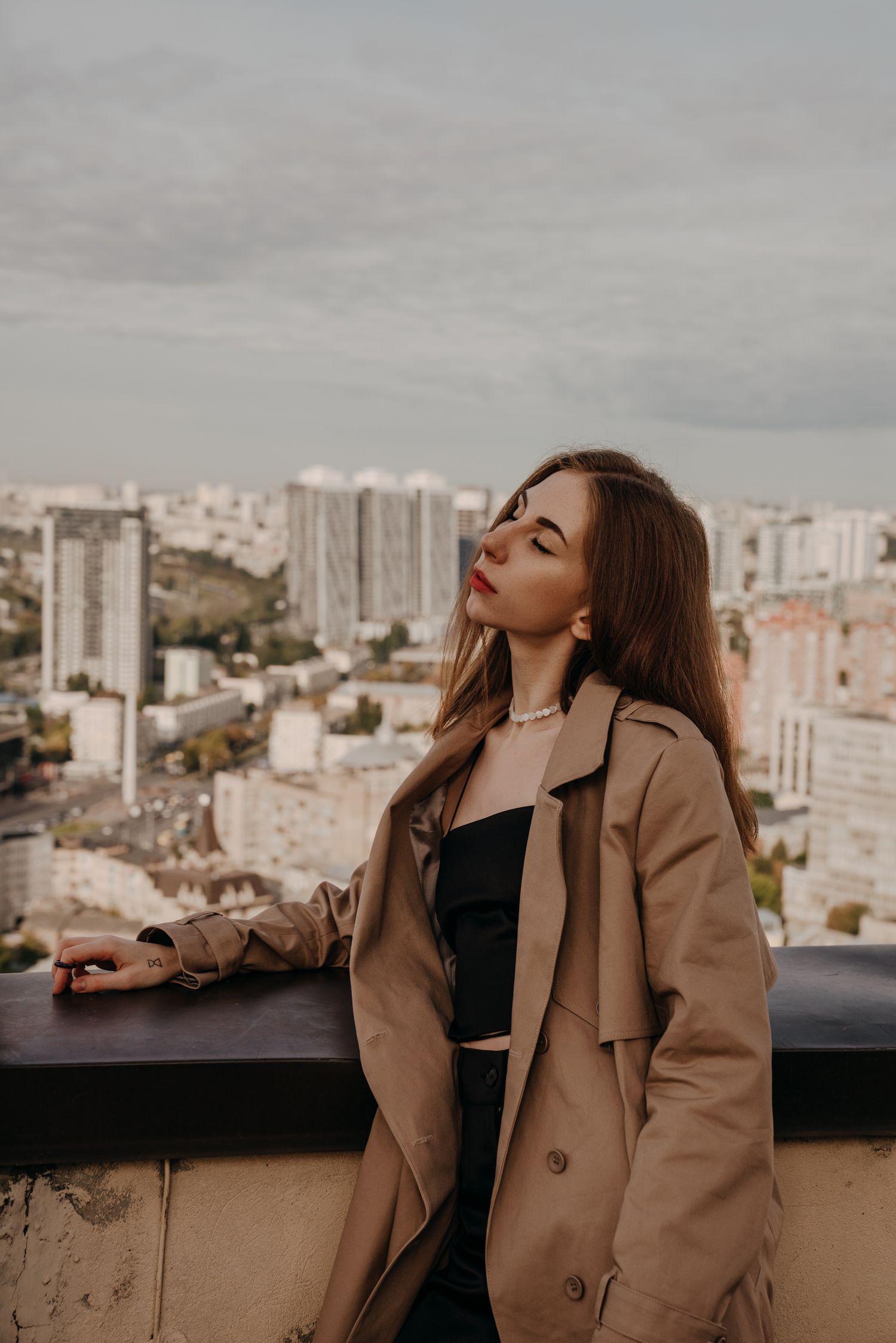 Young woman in beige trench coat poses on rooftop overlooking a city skyline on a cloudy day.