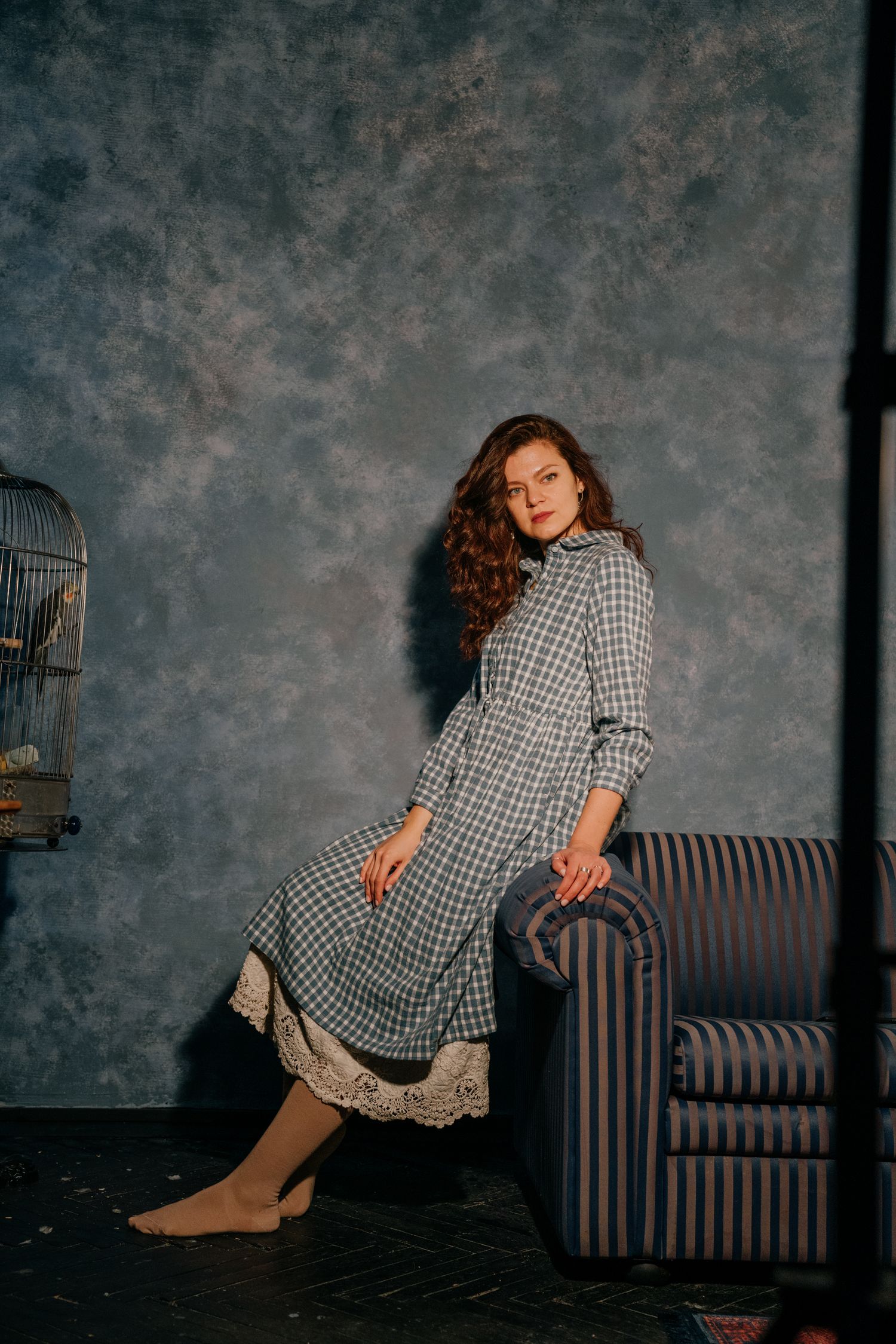 Woman in plaid midi dress with lace trim poses beside striped armchair against gray textured studio wall.